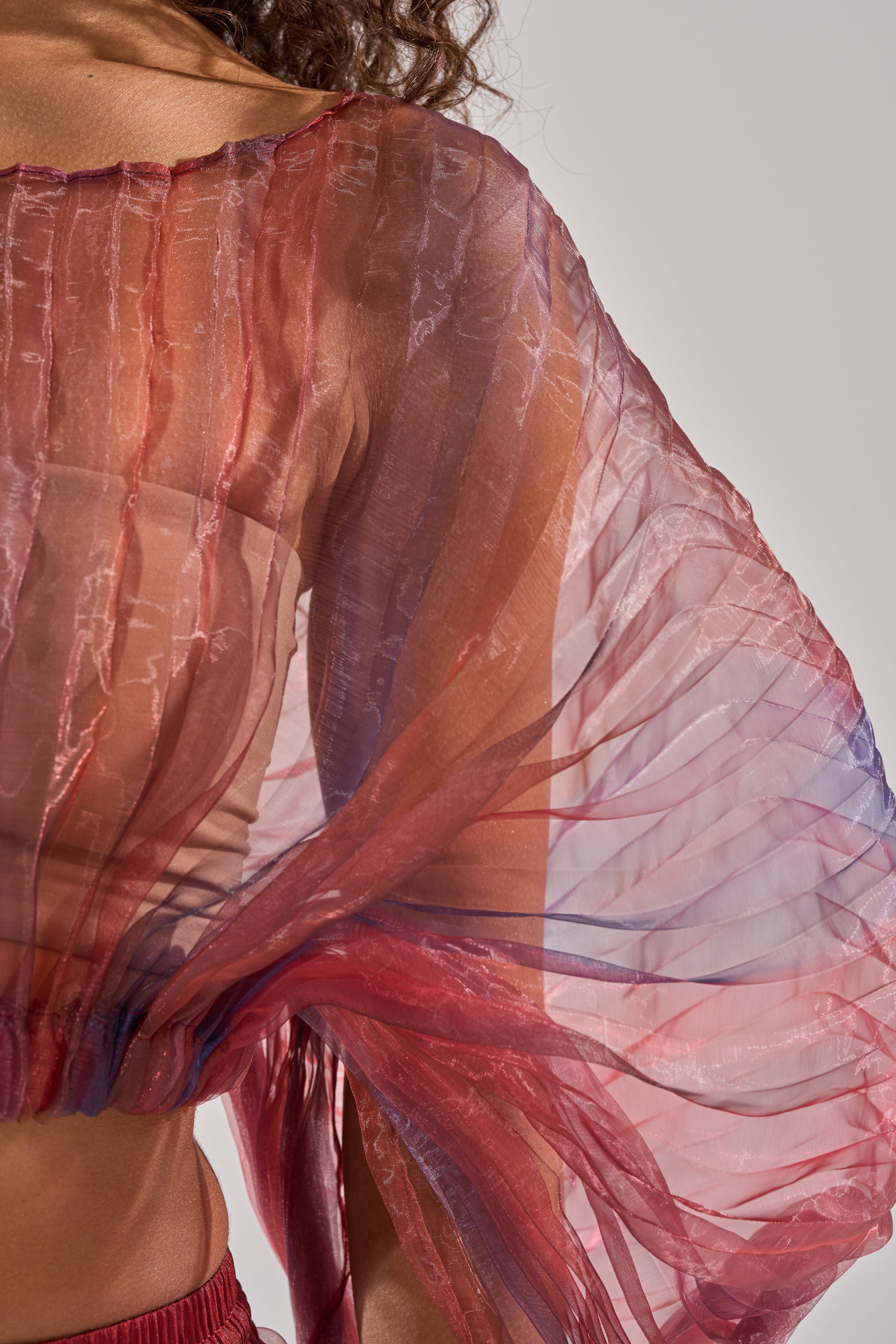A close-up of someone wearing the LETS LIVE IN THE MOMENT CHIFFON BLOUSE, featuring sheer multicolor chiffon with textured vertical lines and delicate accordion pleats, revealing the arm and shoulder through its lightweight, translucent fabric. 8
