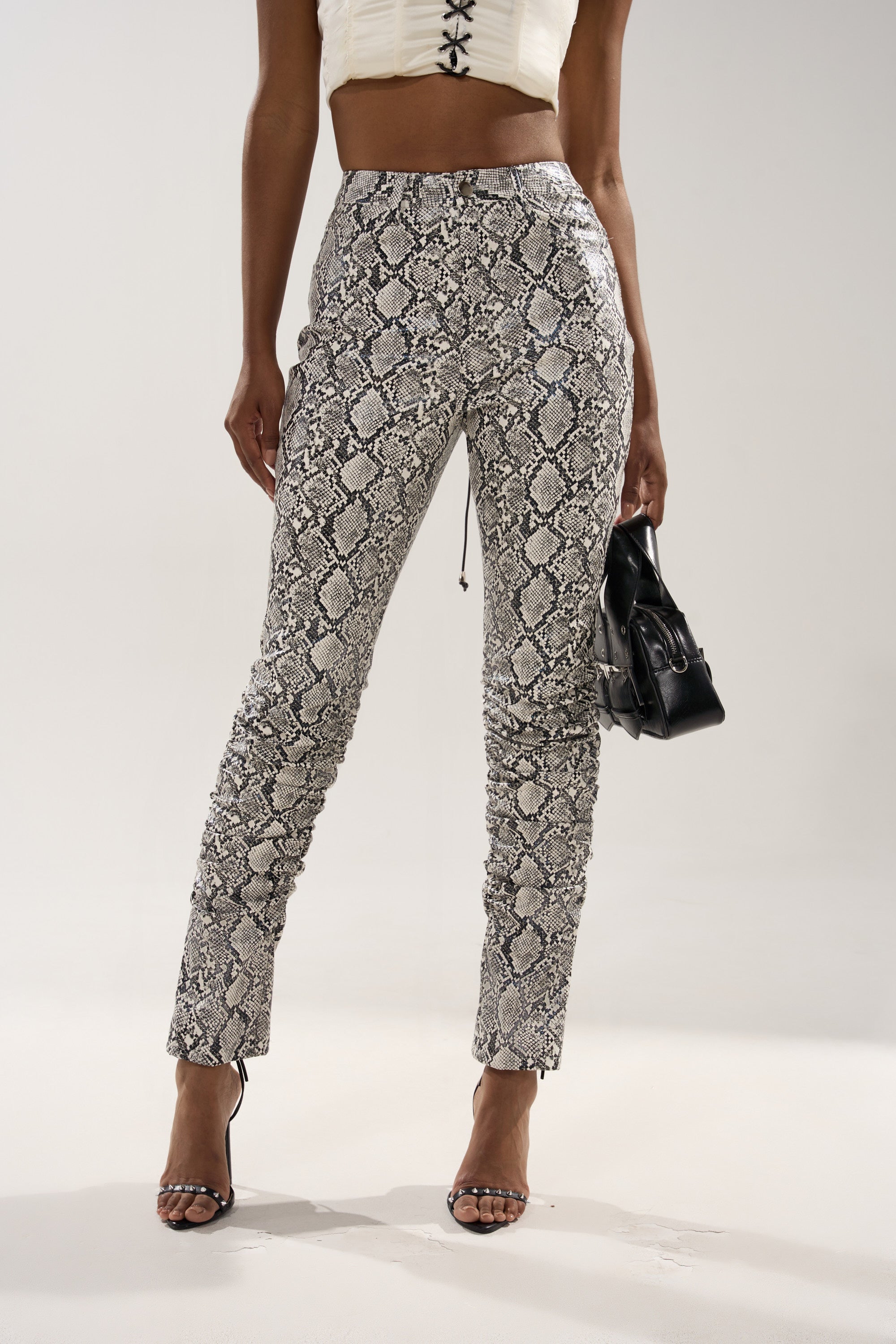 A woman poses against a plain background in the SNEAKY LITTLE SNAKE TROUSER WITH LACEUP ON BUTT, featuring a high-rise faux snakeskin design, paired with a white crop top, black strappy heels, and a small black handbag.