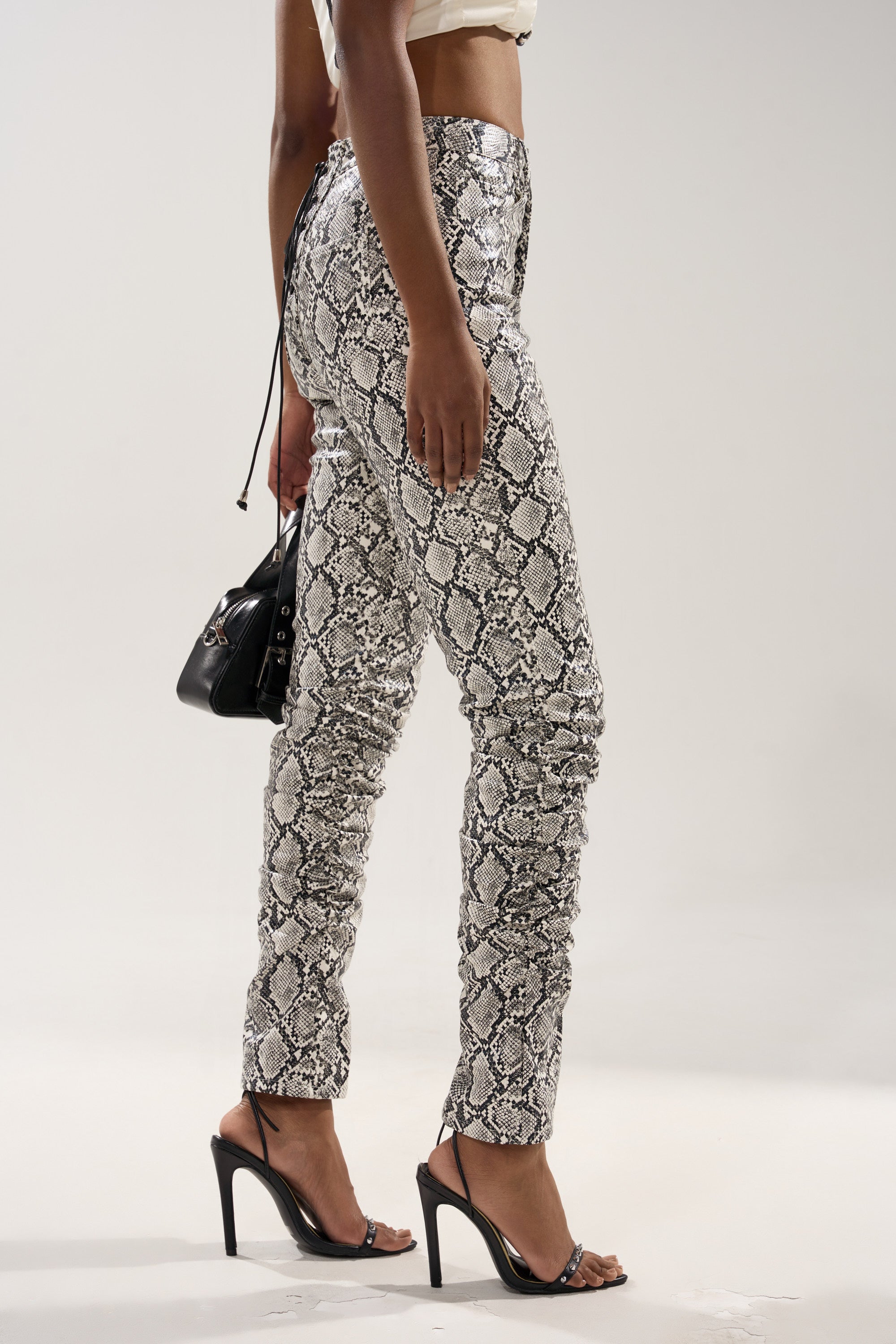 A person wears the Sneaky Little Snake Trouser with lace-up on the butt, featuring a high-rise snakeskin print, paired with black strappy high heels and a small black handbag, shown in profile against a plain background.