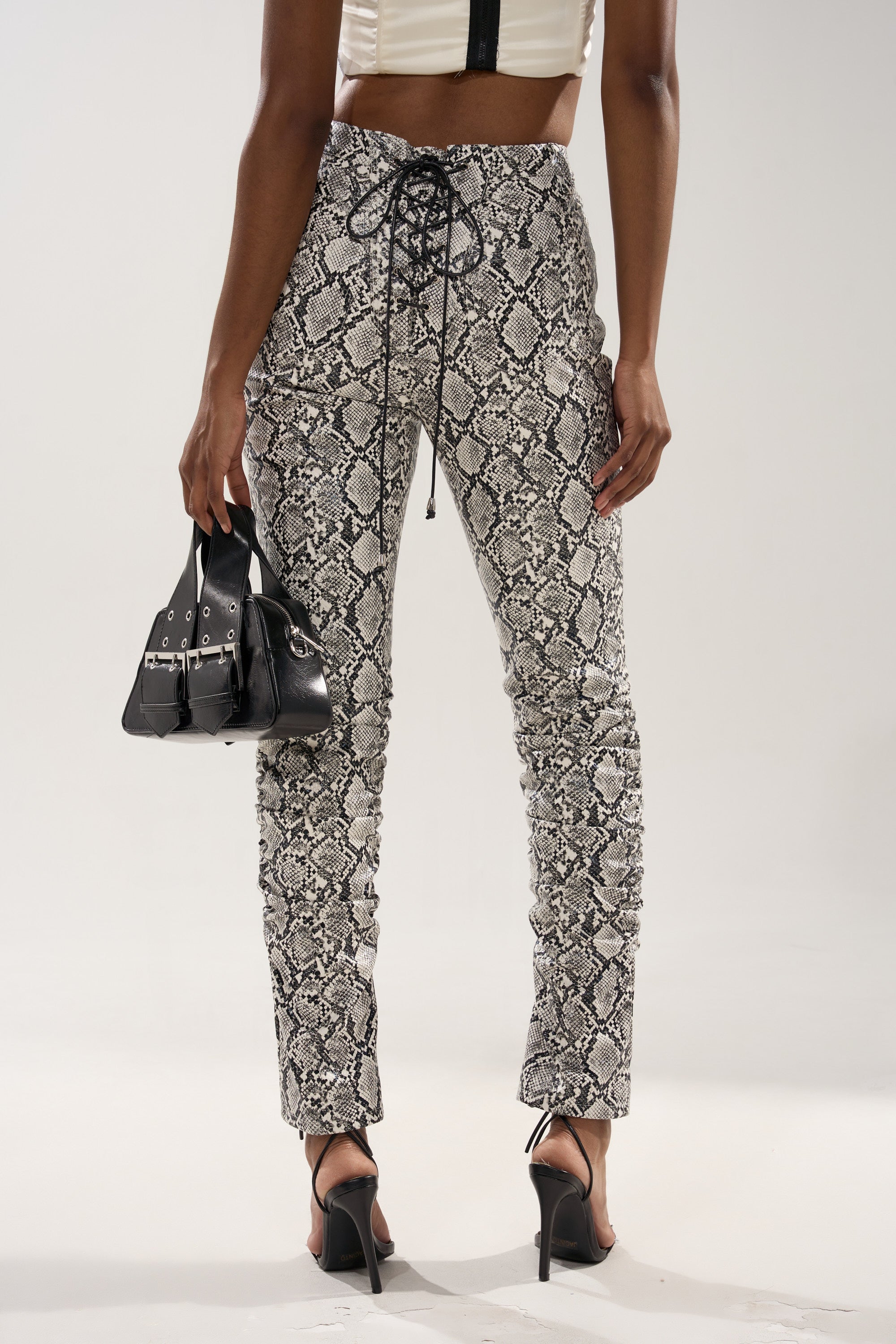 A person stands facing away, wearing the Sneaky Little Snake Trouser with lace-up on the butt, black high-heeled sandals, and a white crop top. They hold a small black handbag, set against a plain, light-colored background.
