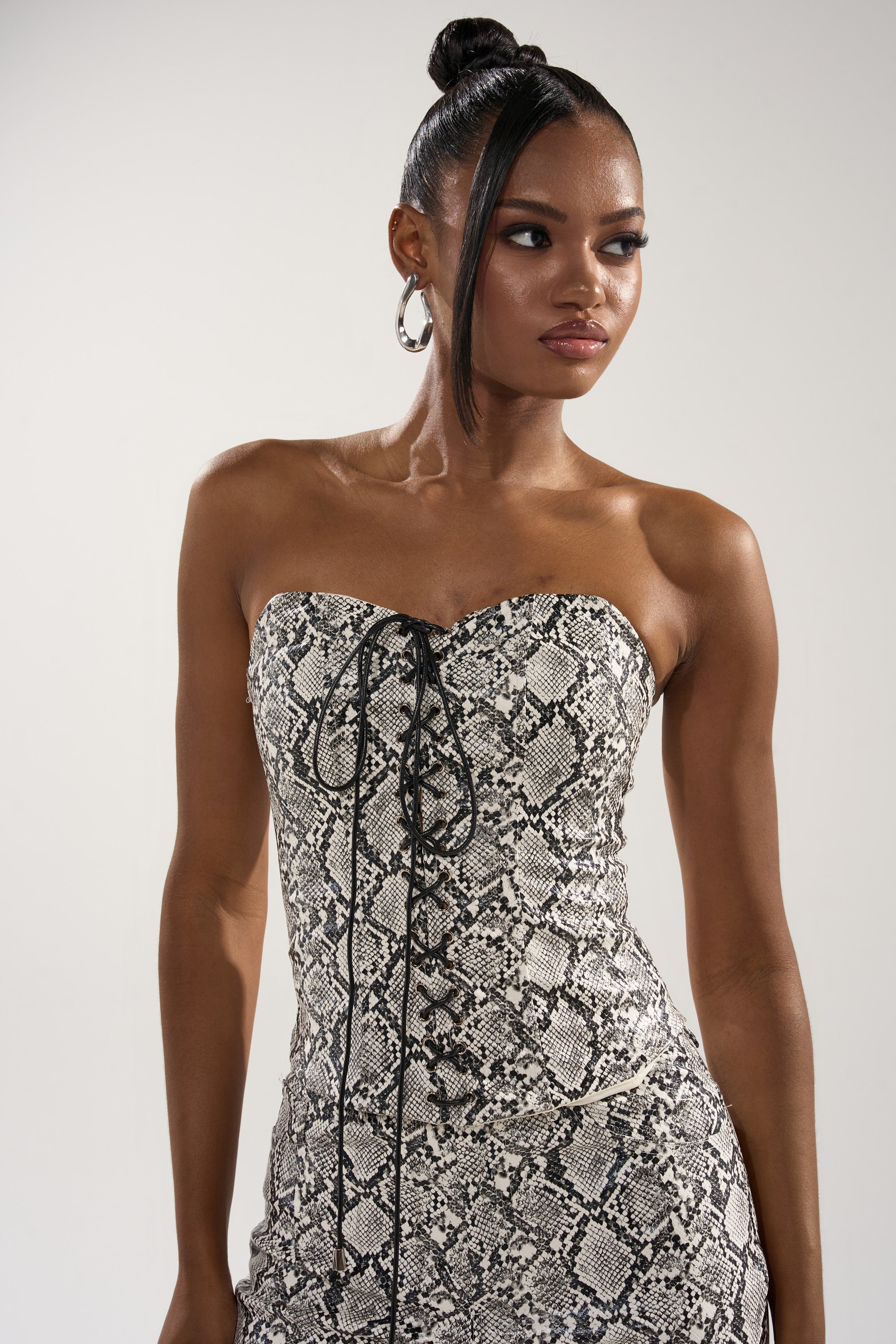 A woman with long hair in a sleek bun models the SNEAKY LITTLE SNAKE CORSET, a strapless black-and-white faux snakeskin dress with a lace-up front, posing confidently against a light background and looking to her left.