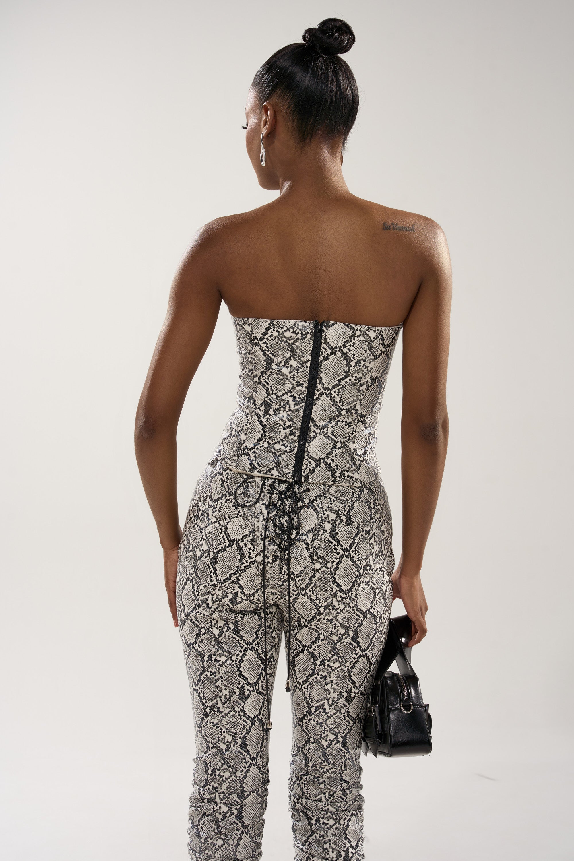 A woman with her hair in a bun stands with her back to the camera, wearing the strapless SNEAKY LITTLE SNAKE CORSET and holding a small black handbag.