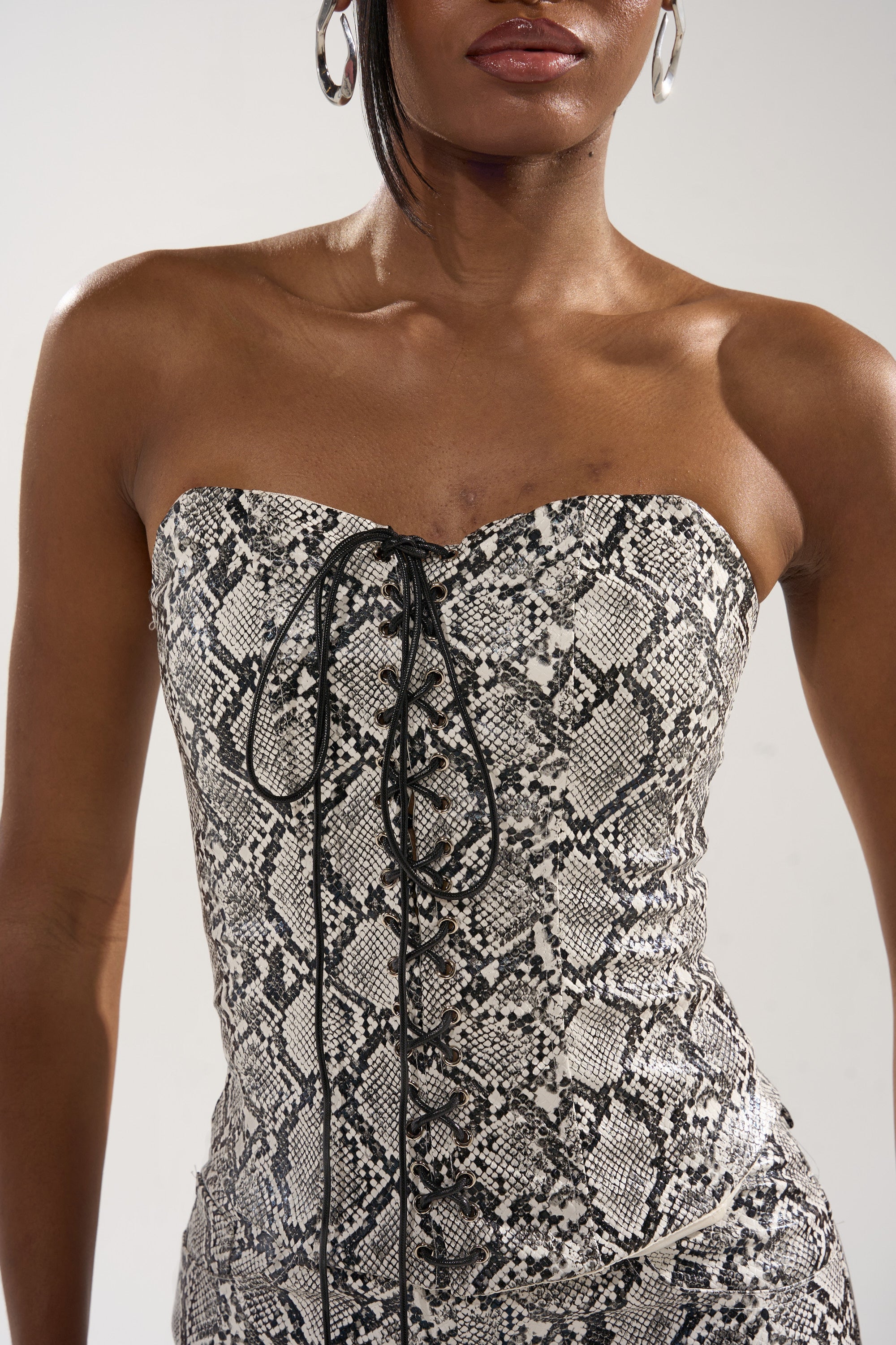 A woman models the SNEAKY LITTLE SNAKE CORSET, a strapless black and white faux snakeskin dress with a lace-up front, short hair, and large hoop earrings, photographed from shoulders to mid-thigh.
