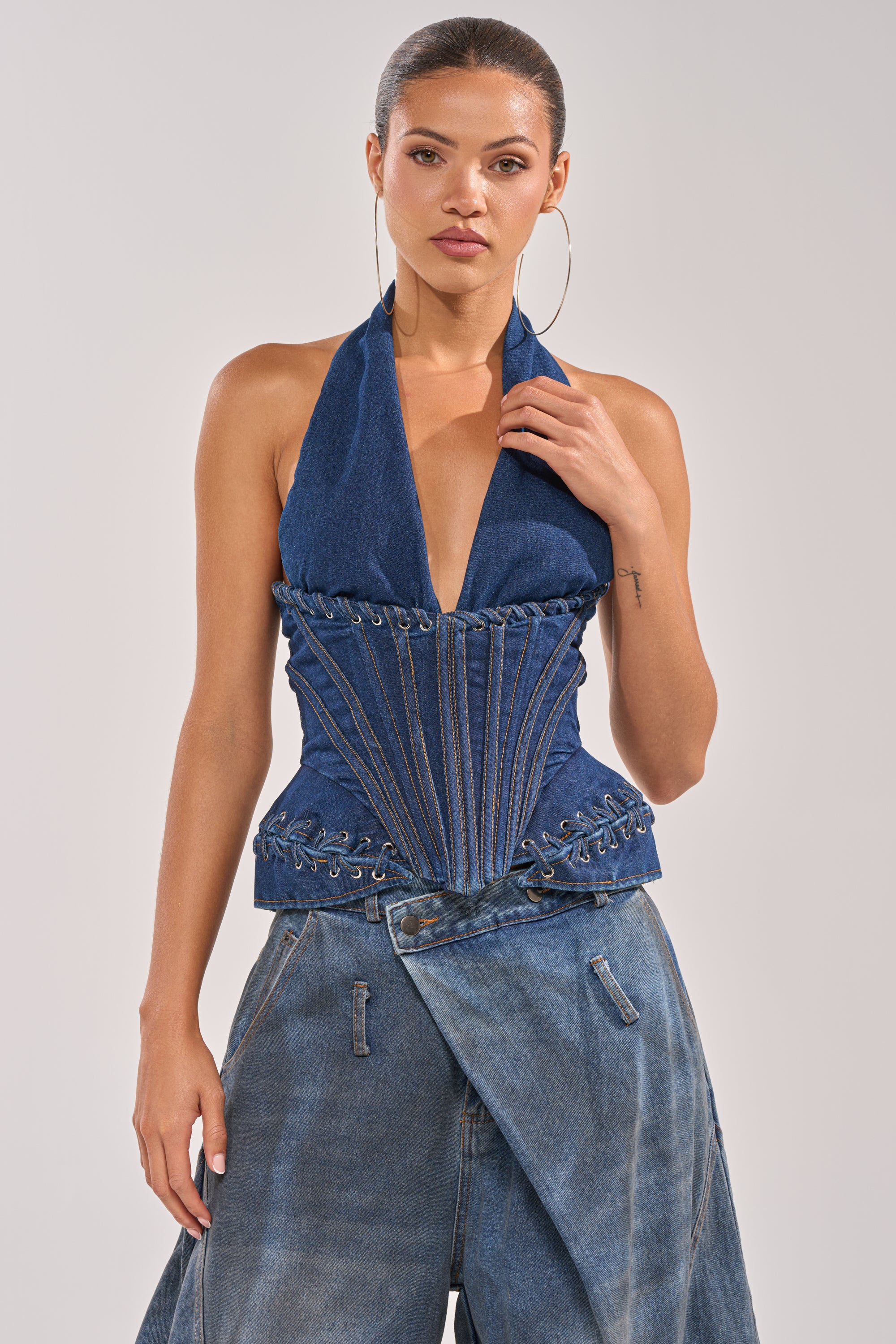 A woman stands facing forward against a plain light background, wearing the NOT MY PROBLEM DENIM CORSET with a deep V-neck halter style and corset lacing, paired with baggy asymmetrical denim pants. 0