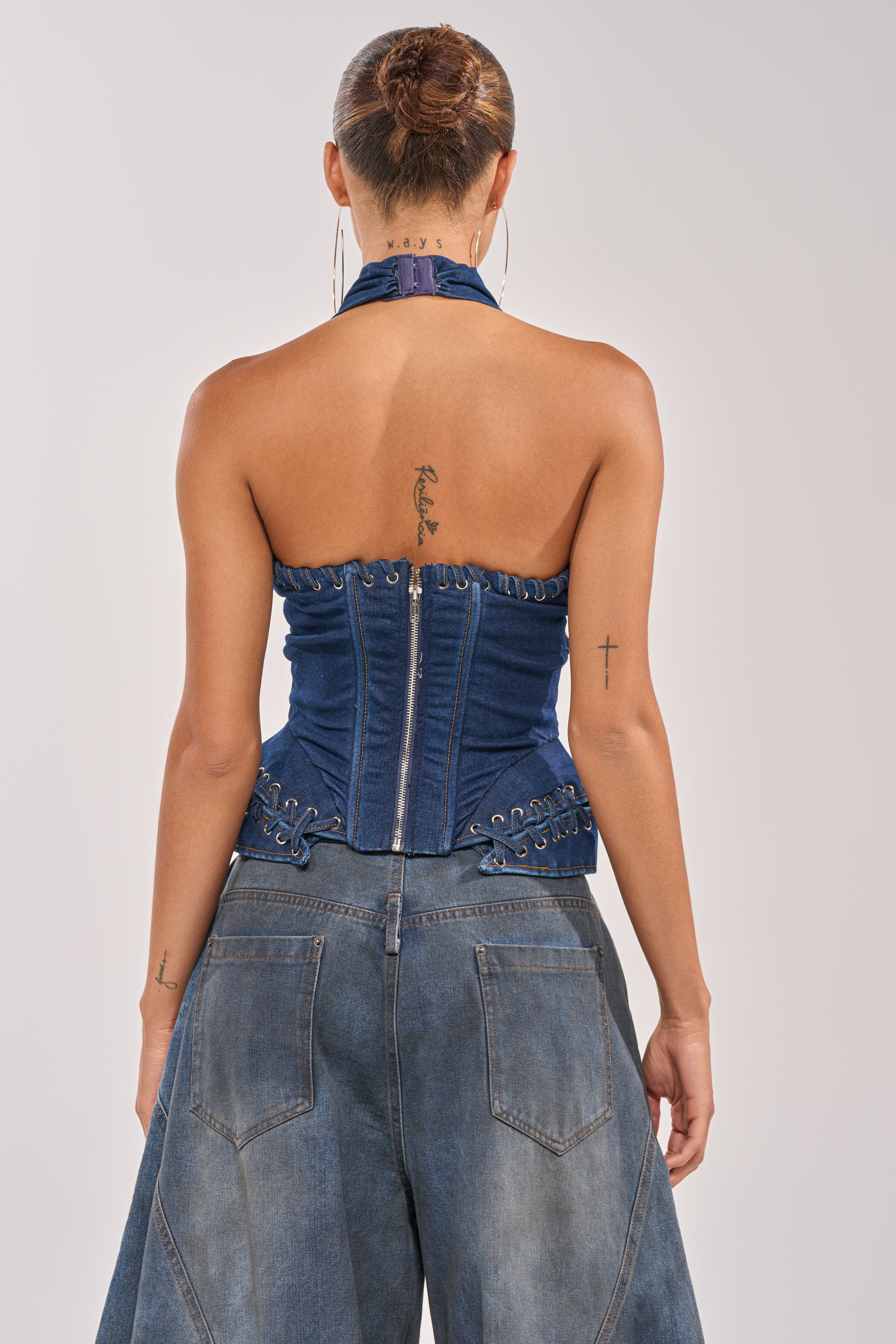 A person with their hair in a bun wears the NOT MY PROBLEM DENIM CORSET and wide-legged jeans, seen from behind. Tattoos are visible on their neck, upper back, and right arm. 3