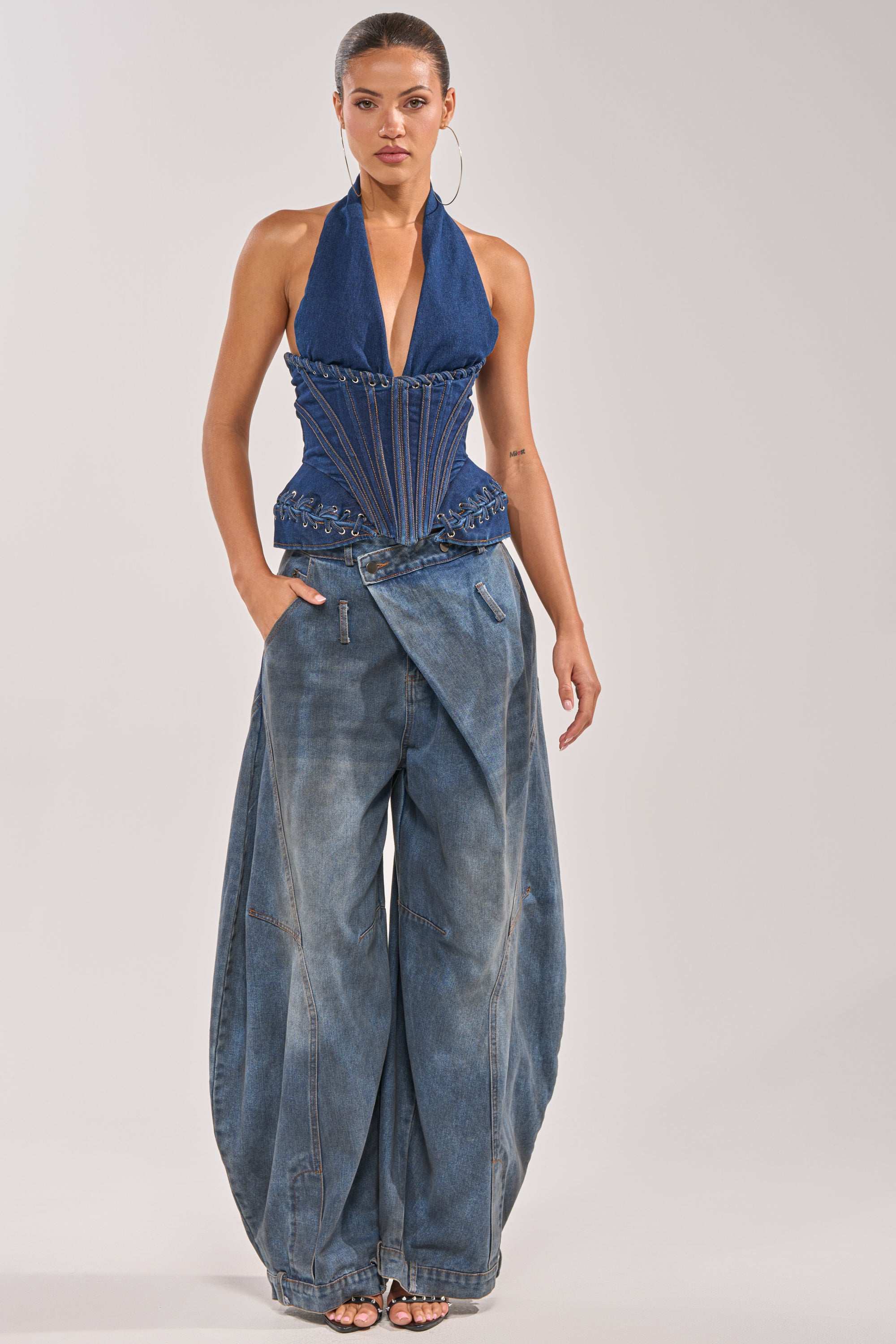 A woman wears the NOT MY PROBLEM DENIM CORSET, a deep blue, western-inspired halter-neck top with decorative stitching, paired with wide, high-waisted baggy denim pants. She stands against a plain background with her hair pulled back and hoop earrings on. 4