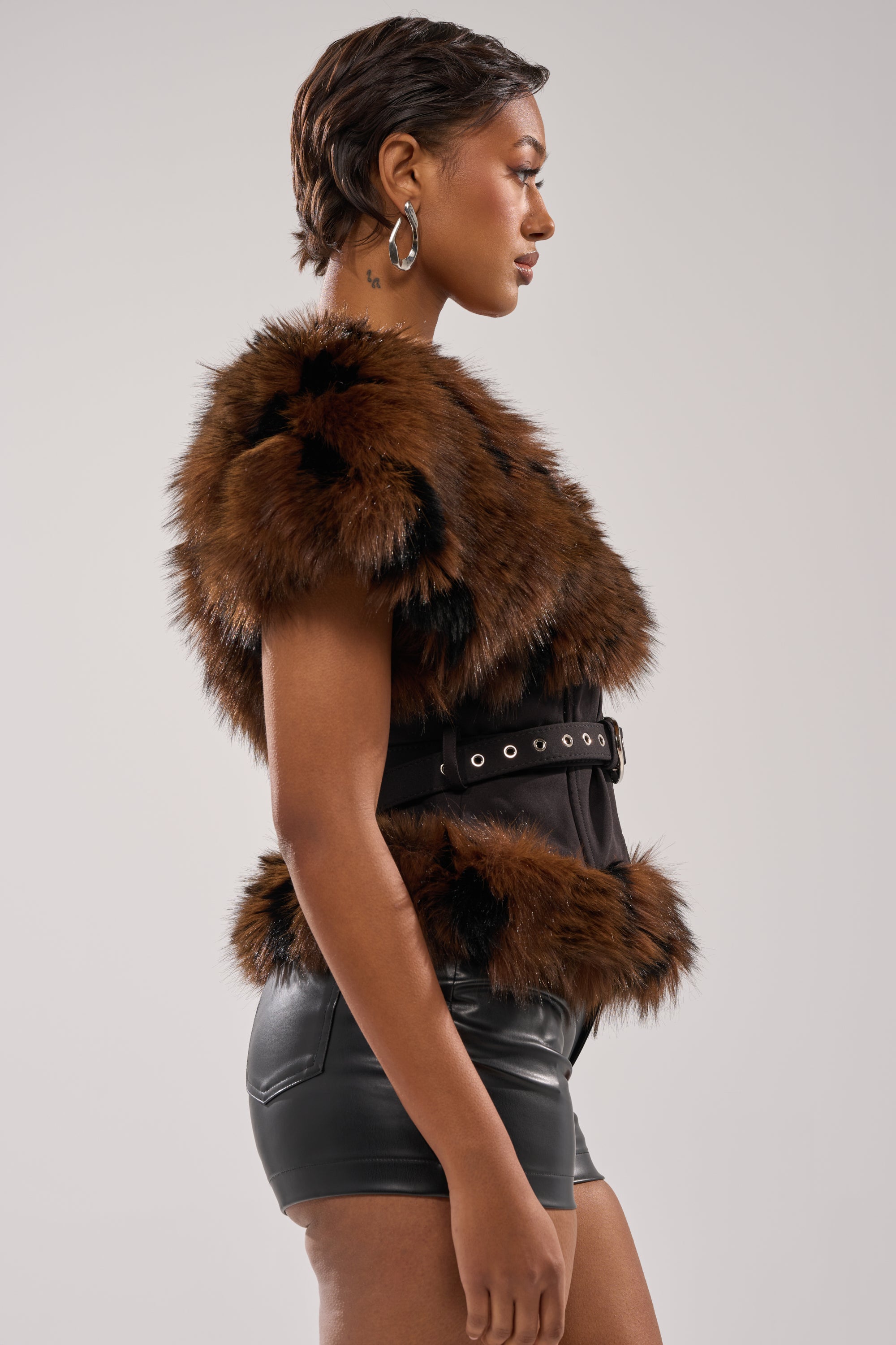 A woman poses in profile against a plain light background, wearing the FEELING WARM FUR BLOUSE—styled winter-chic with leather shorts, a black adjustable belt top, and hoop earrings. 3