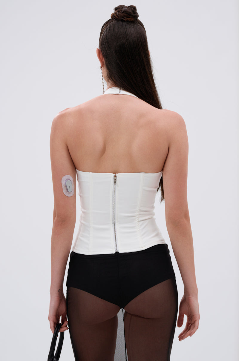 A woman with long dark hair is shown from the back wearing the LOVE IS REAL BOW HALTER TOP and black sheer pants. She has a medical device on her upper left arm and holds a small black bag. 3