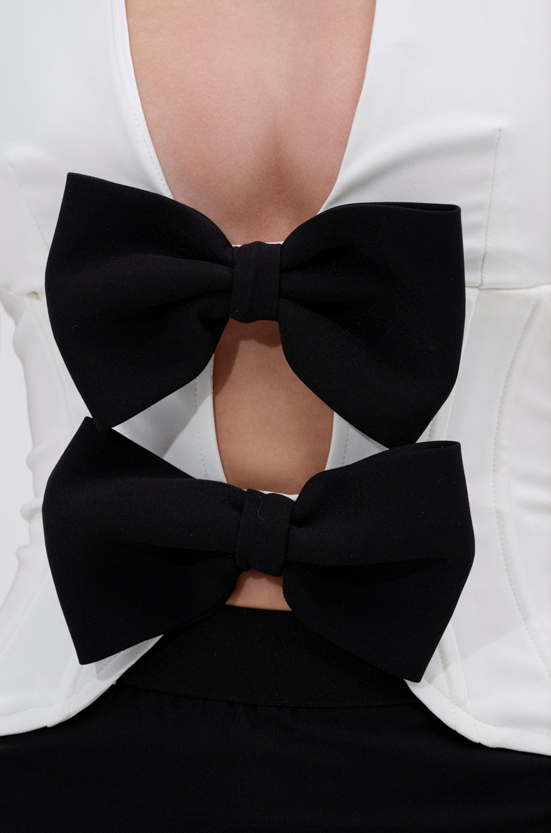 A close-up of the LOVE IS REAL BOW HALTER TOP, a white halter crop with a deep plunging neckline and two bold black bows, revealing sexy midriff cut-outs. Styled with a sleek black bottom. 6