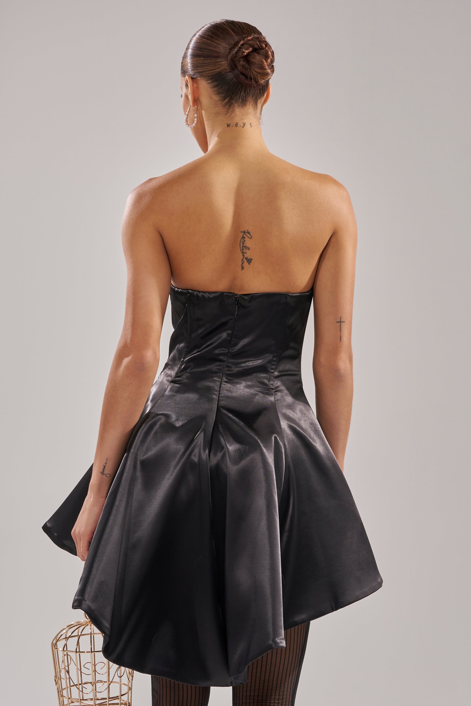 A woman with tattoos on her upper back and arms is shown from behind, wearing the PRETTY PLEASE SATIN CORSET IN BLACK— a strapless, sweetheart neckline dress with a flared skirt— her hair styled in a sleek bun. 3