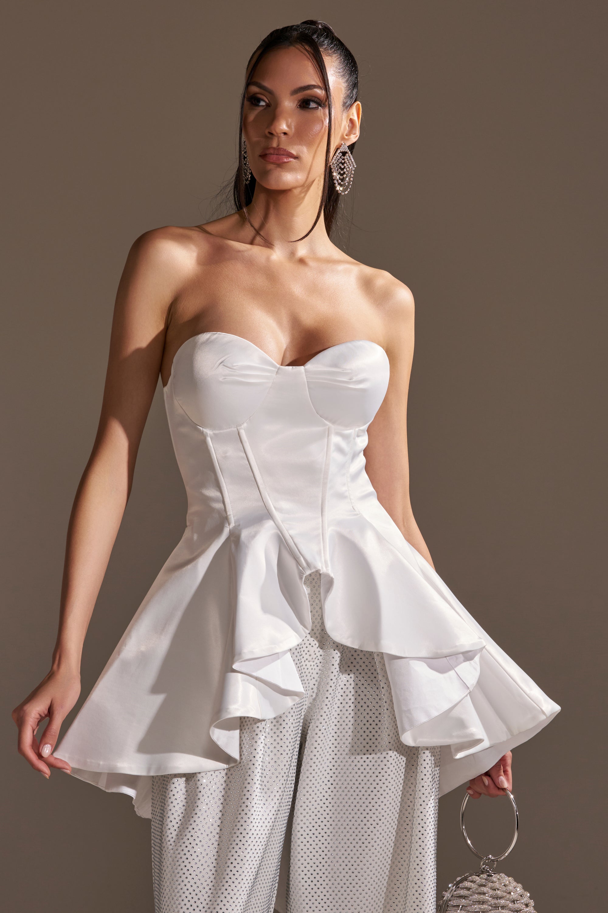 A woman poses against a plain background wearing the PRETTY PLEASE SATIN CORSET with flared ruffles, wide-leg textured pants, large hoop earrings, and a small silver handbag.