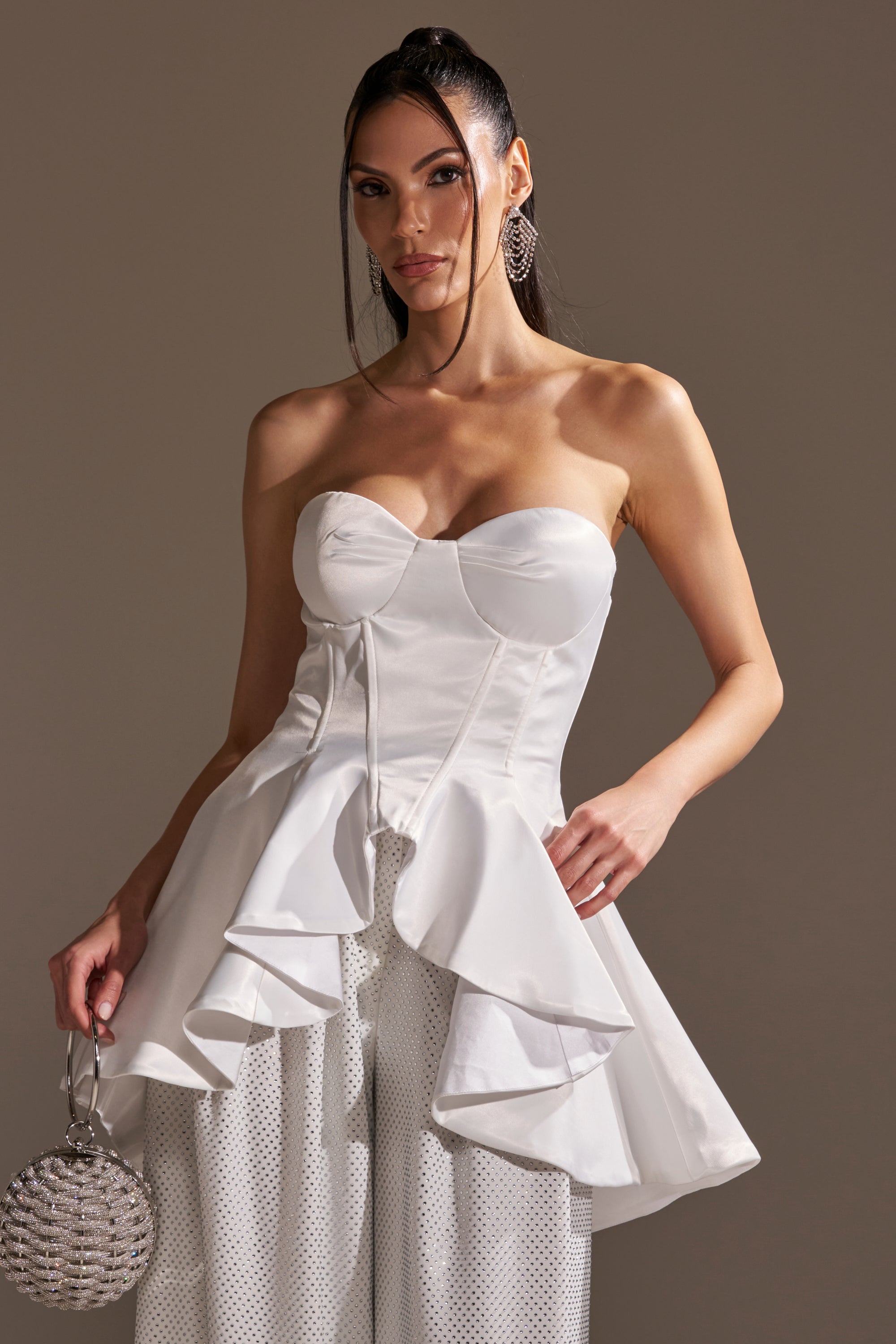 A woman stands against a neutral background wearing the PRETTY PLEASE SATIN CORSET—a strapless, sweetheart neckline white peplum top with a high-low hem—paired with a round rhinestone handbag and large sparkling earrings.