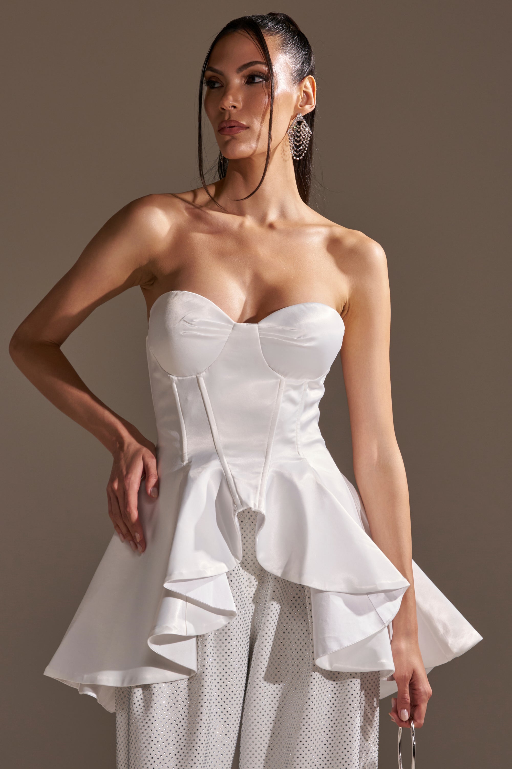 A woman stands against a neutral background, wearing the PRETTY PLEASE SATIN CORSET with white textured pants. She looks slightly to the side, hand on her hip, and her long dark hair is styled in a sleek ponytail.