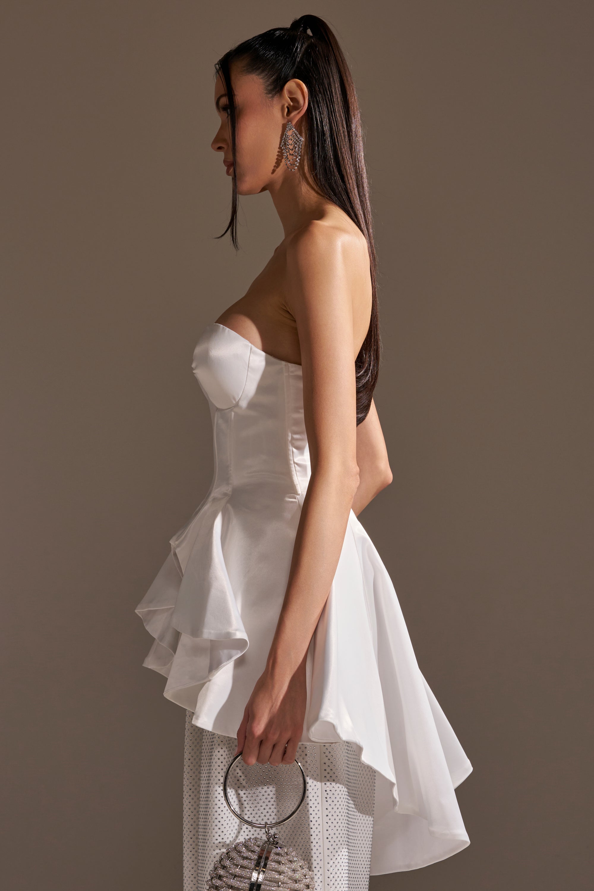 A woman in profile wears the PRETTY PLEASE SATIN CORSET with a sweetheart neckline, flared peplum hem, white textured pants, and a small handbag. Her long, straight hair is styled in a high ponytail.