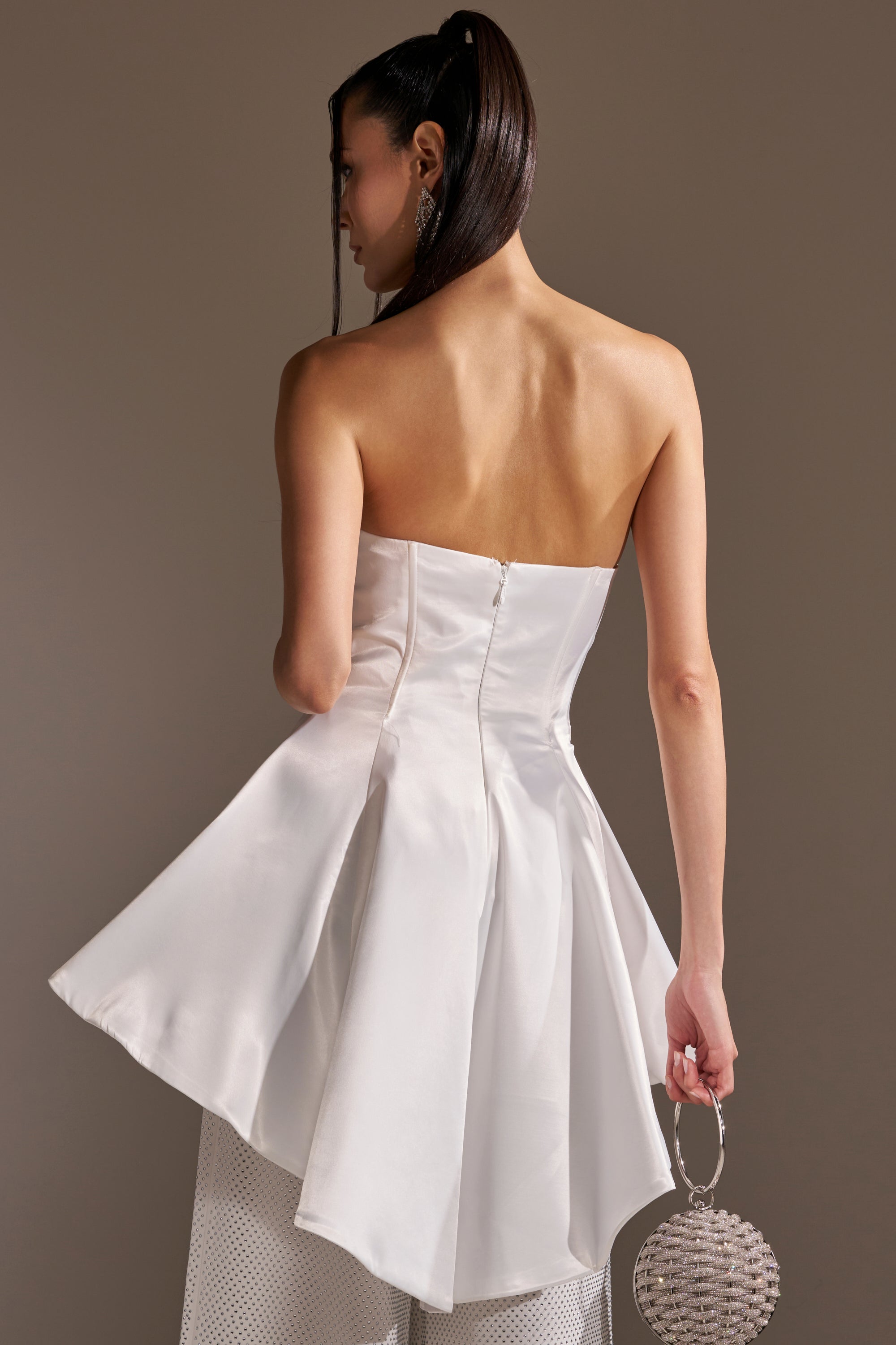 A woman with long dark hair in a ponytail wears the PRETTY PLEASE SATIN CORSET, a white strapless sweetheart neckline top with a high-low peplum hem, paired with white mesh pants and holding a round silver beaded handbag against a neutral background.