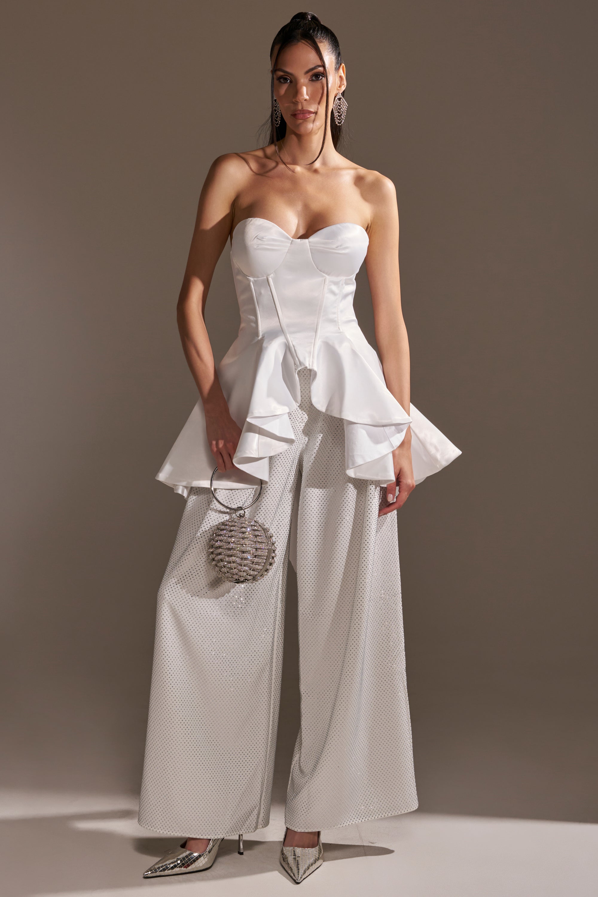A woman poses against a neutral background wearing the PRETTY PLEASE SATIN CORSET with wide-leg textured white pants, a round silver purse, metallic pointed-toe shoes, sleeked-back hair, and statement earrings.