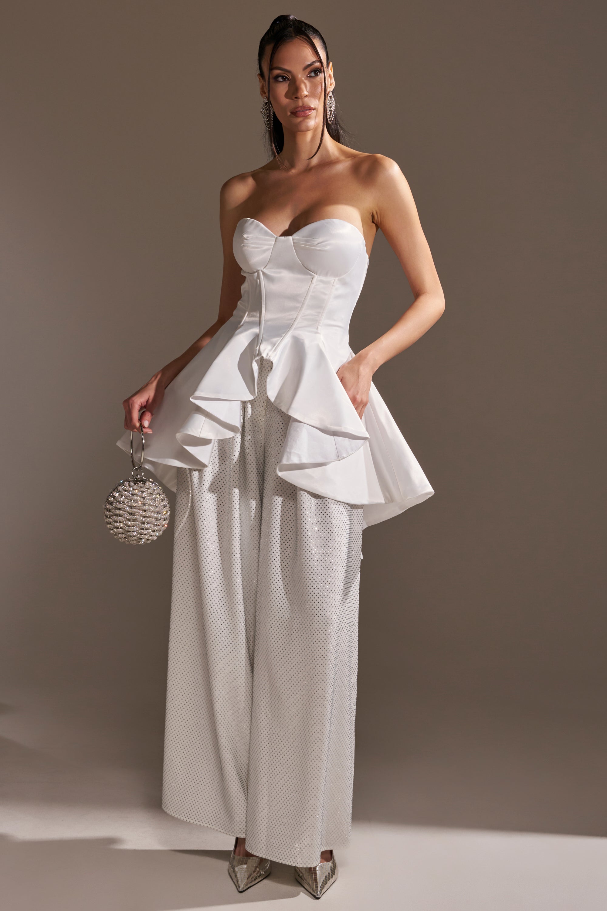 A woman wears the PRETTY PLEASE SATIN CORSET, a strapless white peplum top with structured ruffles, paired with wide-leg textured pants, a silver beaded handbag, and pointed metallic heels against a neutral background.