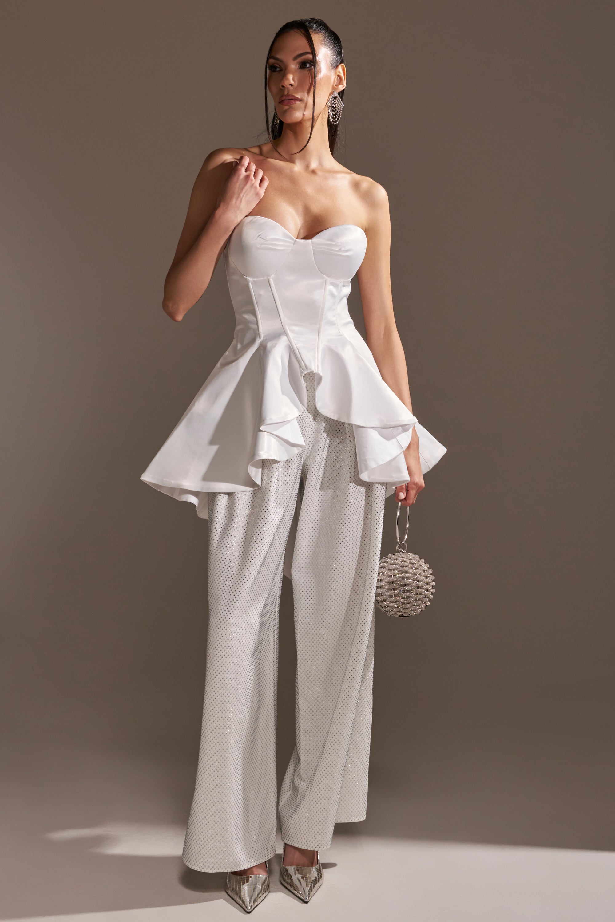 A woman wears the PRETTY PLEASE SATIN CORSET, a strapless, sweetheart neckline top with peplum ruffles, paired with wide-leg white pants, a round silver purse, metallic pointed heels, and a sleek updo.