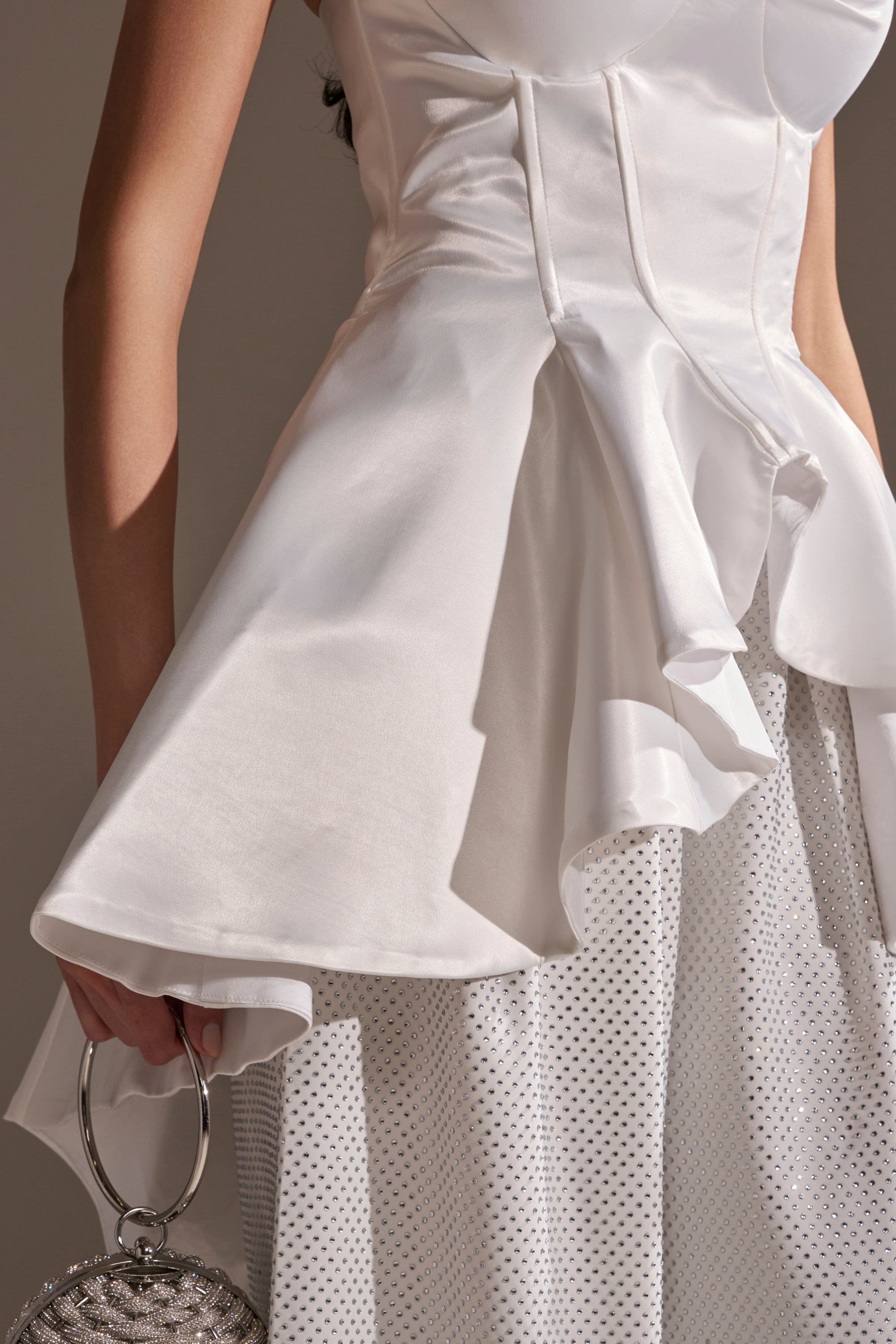 Wearing the PRETTY PLEASE SATIN CORSET, a white structured top with a flared high-low peplum hem, paired with a white polka dot skirt and holding a silver round handbag with a large ring handle.