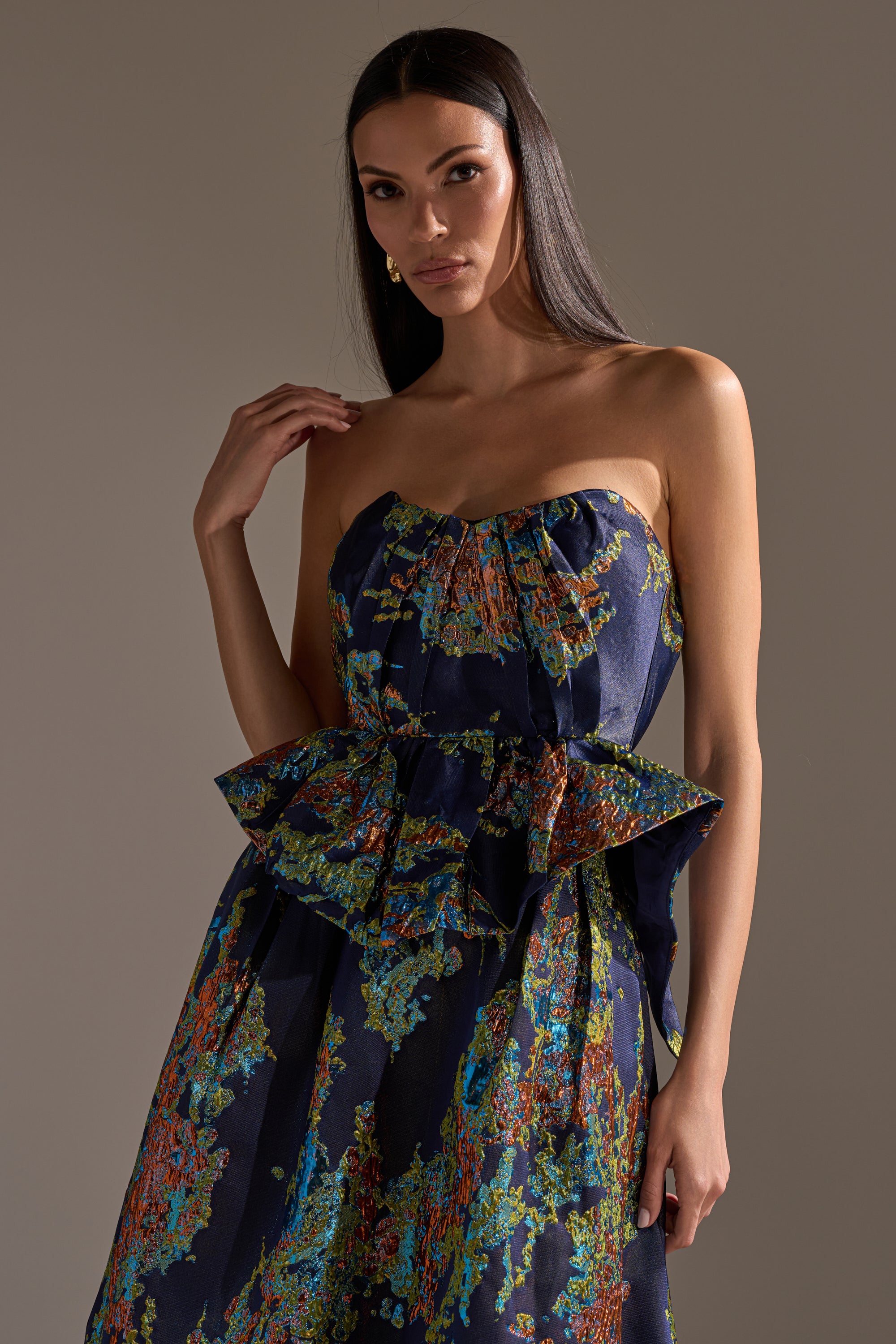 A woman with long dark hair models the TRANSCENDENT FLORAL BROCADE CORSET TOP, a strapless dark piece with a vibrant abstract floral pattern and peplum waist detail, posing with one hand on her shoulder against a neutral background.