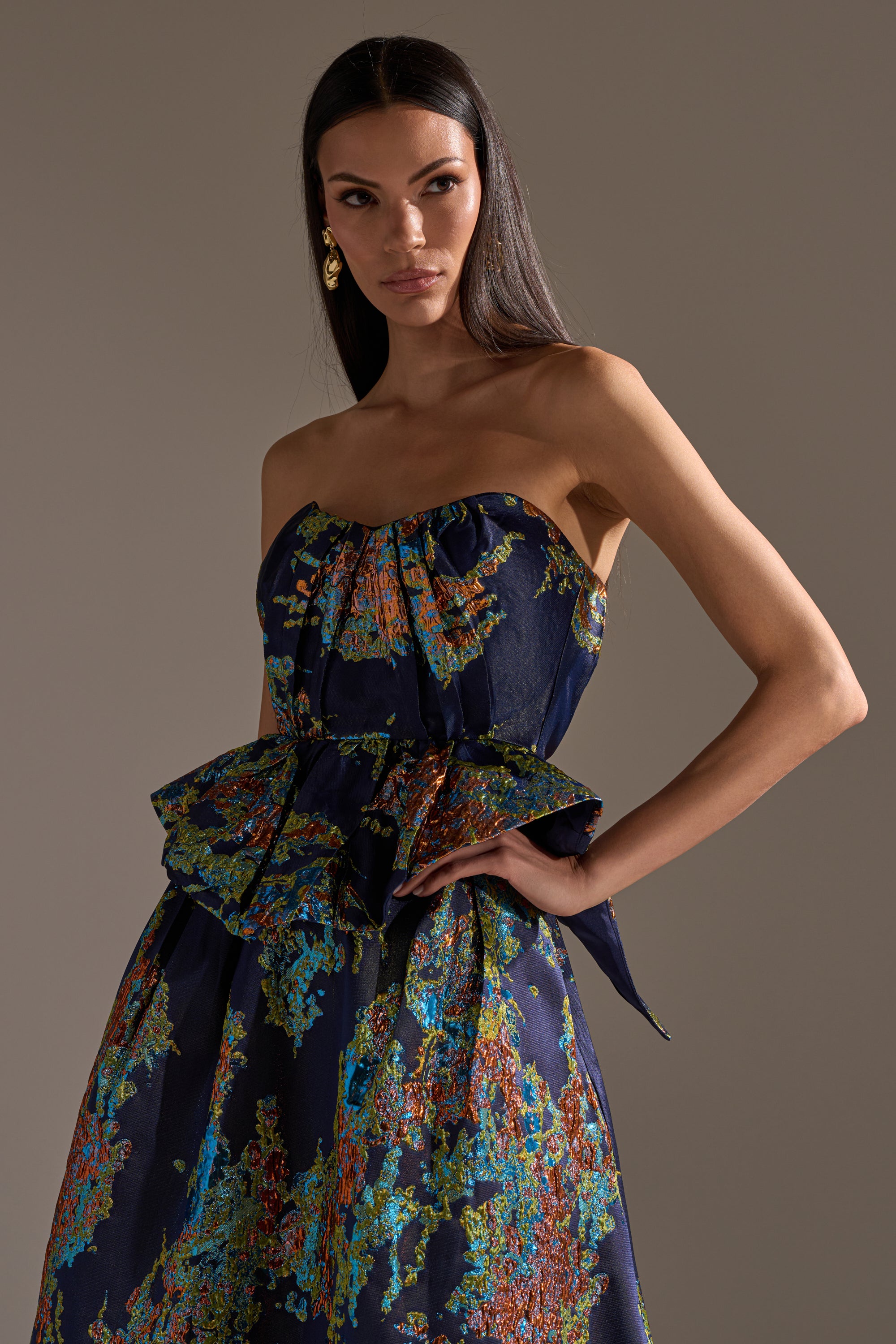 A woman poses with one hand on her hip, wearing the TRANSCENDENT FLORAL BROCADE CORSET TOP featuring a strapless sweetheart neckline, colorful abstract floral pattern, and ruffled peplum detail against a neutral background.