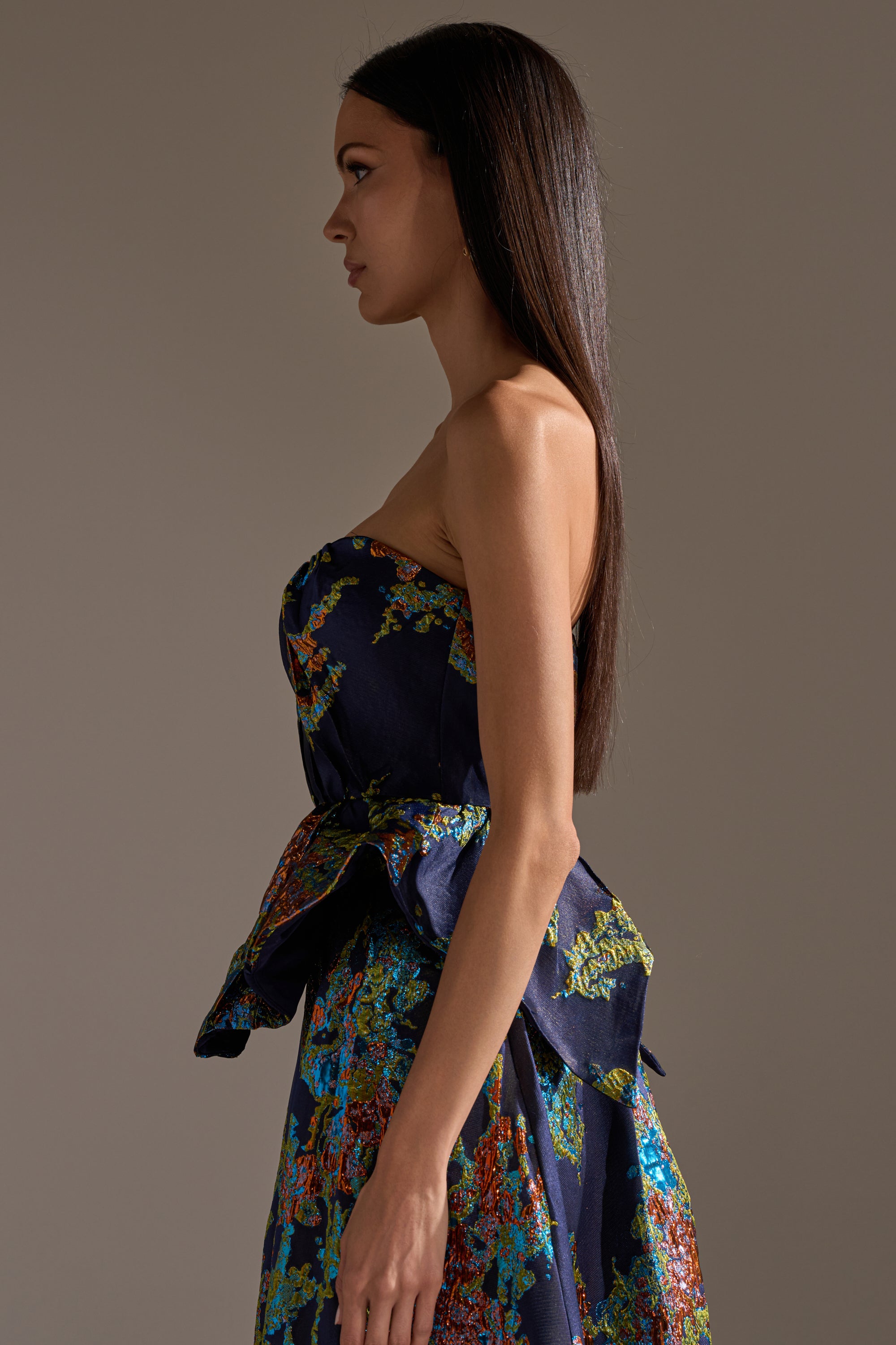 A woman with straight, long dark hair stands in profile against a neutral background, wearing the TRANSCENDENT FLORAL BROCADE CORSET TOP styled as a navy blue dress with colorful patterns and a peplum detail at the waist.