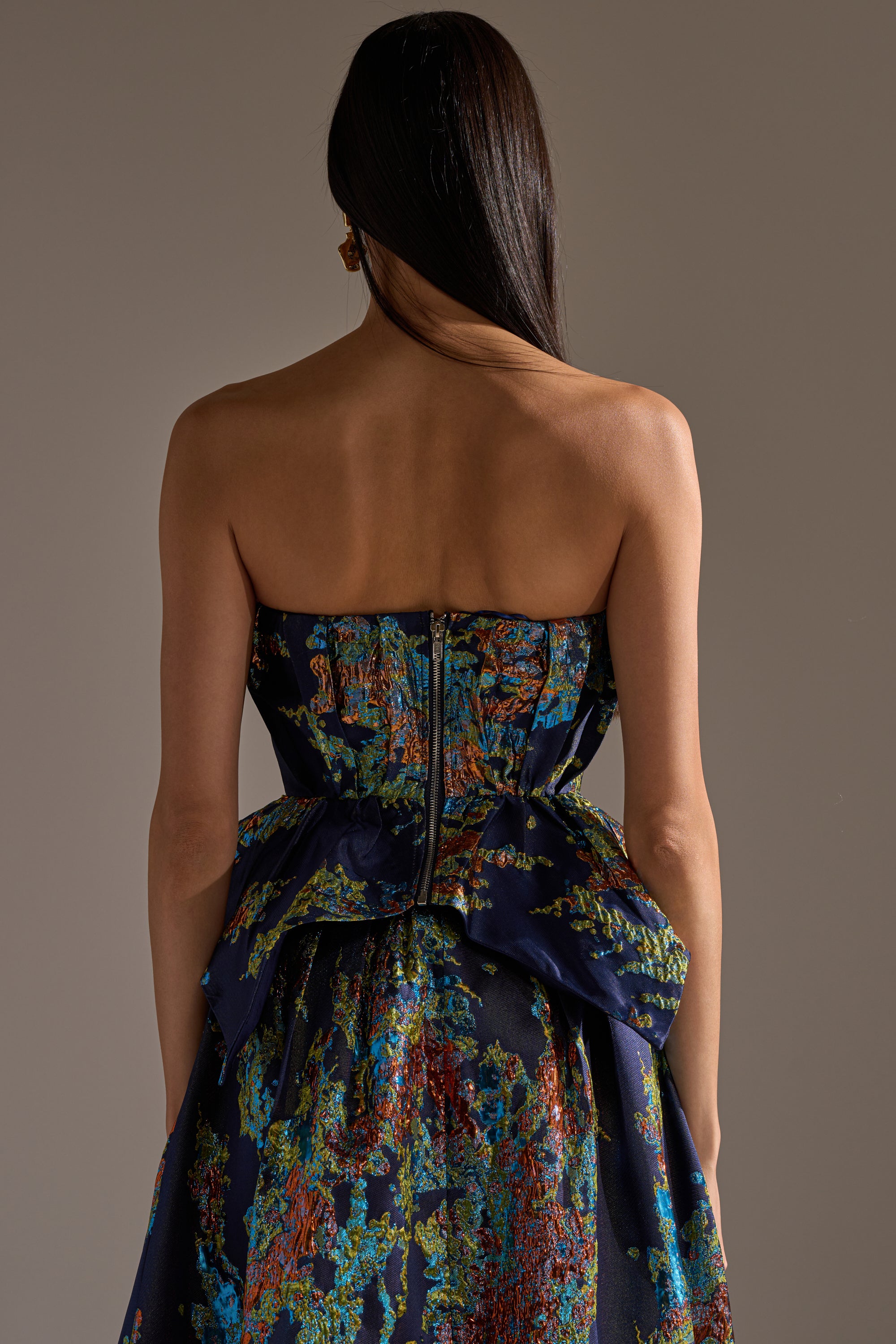 A woman with long, straight hair is seen from behind wearing the TRANSCENDENT FLORAL BROCADE CORSET TOP, a strapless dark blue piece with a peplum waist and colorful floral pattern, set against a plain background.