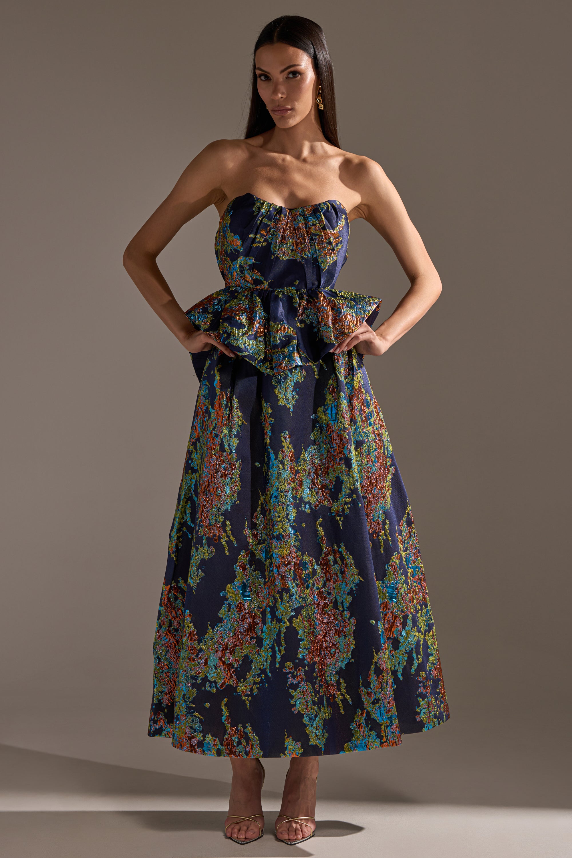 A woman models the TRANSCENDENT FLORAL BROCADE CORSET TOP—strapless, with a colorful abstract floral pattern and ruffled peplum waist—posing hands on hips in an ankle-length dress, exuding elegance.