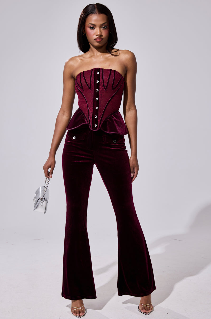 A woman exudes confidence in the Ruby Rose Velvet Trouser, paired with a matching burgundy strapless velvet corset top, silver handbag, and strappy heels, standing against a simple backdrop. 0