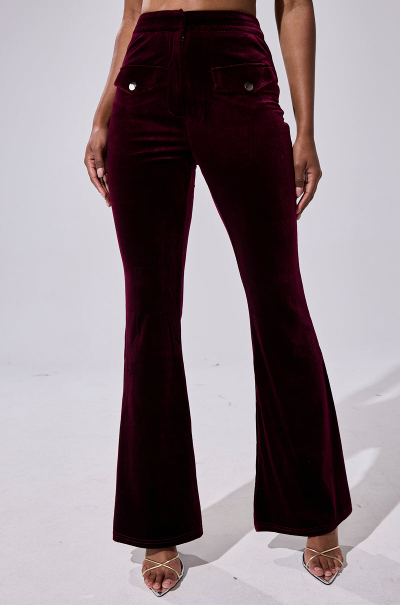 Wearing the RUBY ROSE VELVET TROUSER—high-rise, dark burgundy flared pants with front pockets and silver buttons—paired with open-toe heeled sandals, the bold silhouette stands out against a light background. 1