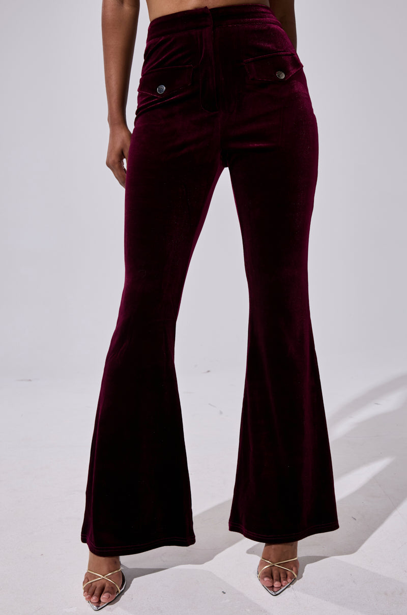 Someone wears the RUBY ROSE VELVET TROUSER, a high-rise, flared-leg pant in dark burgundy velvet with front buttons and pockets, paired with strappy open-toe heels against a plain, light-colored background. 2