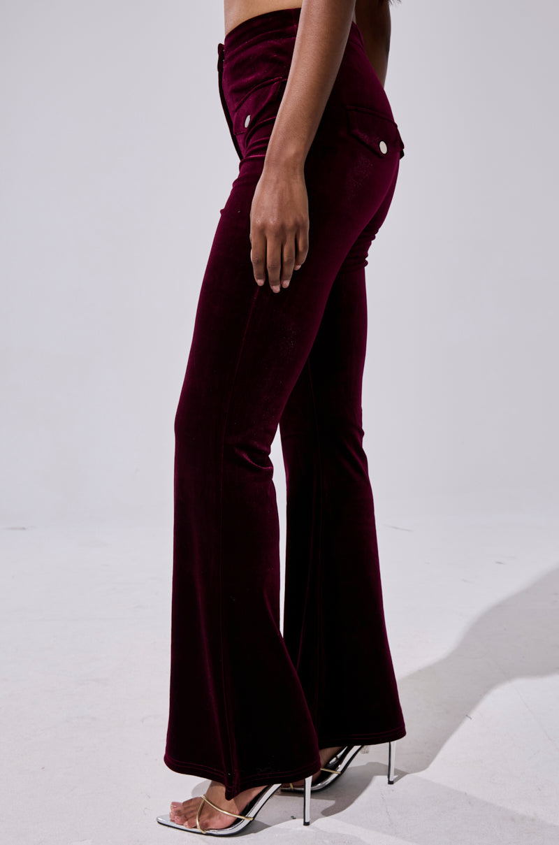 A person stands against a plain light background wearing the RUBY ROSE VELVET TROUSER—high-rise, dark burgundy velvet flared-leg pants with side pockets and silver buttons—paired with open-toe high heels. 3