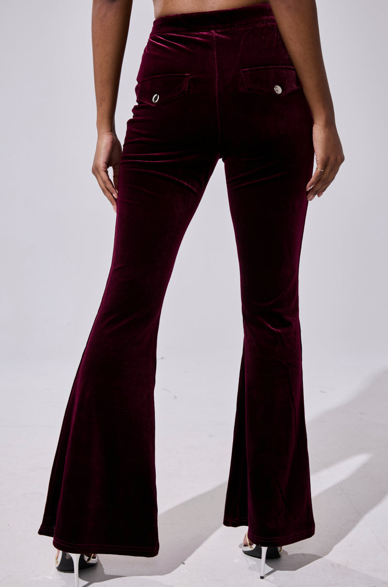 A person stands on a light gray background wearing RUBY ROSE VELVET TROUSER flared-leg, high-rise burgundy velvet pants with buttoned back pockets. The photo, taken from behind, shows the full length and black heels. 4