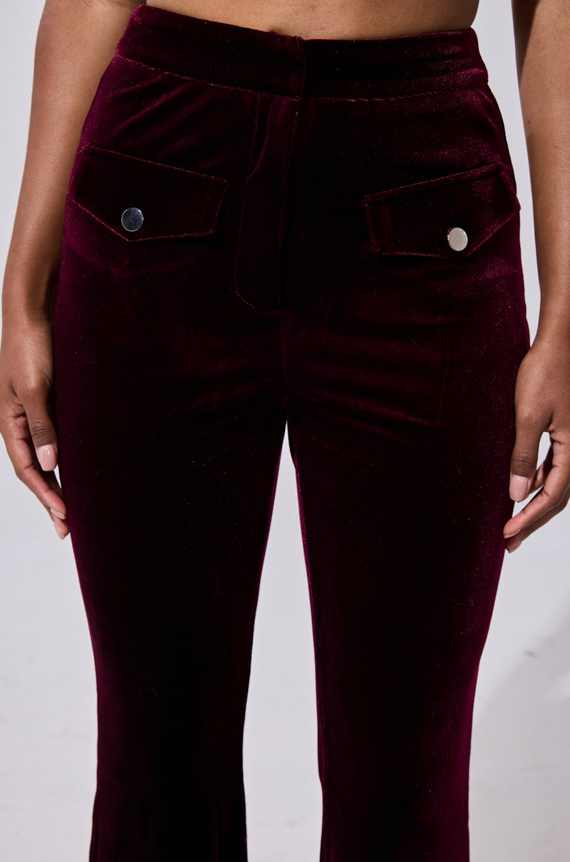 Wearing the RUBY ROSE VELVET TROUSER—high-waisted, dark burgundy, flared-leg velvet pants with front flap pockets and silver buttons—a person stands against a plain light background. 6