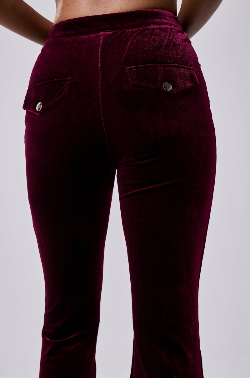 A close-up shows the RUBY ROSE VELVET TROUSER in dark burgundy, featuring a high-rise fit, flared-leg silhouette, and two back flap pockets with metal buttons, set against a plain light gray background. 7