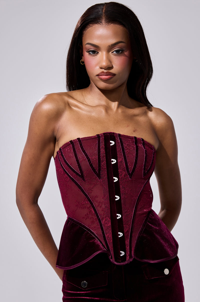 A woman with long dark hair wears the RUBY ROSE CORSET, featuring deep red velvet panels, black accents, and silver eyelets, standing against a plain light gray background with a neutral expression and her hands behind her back. 1