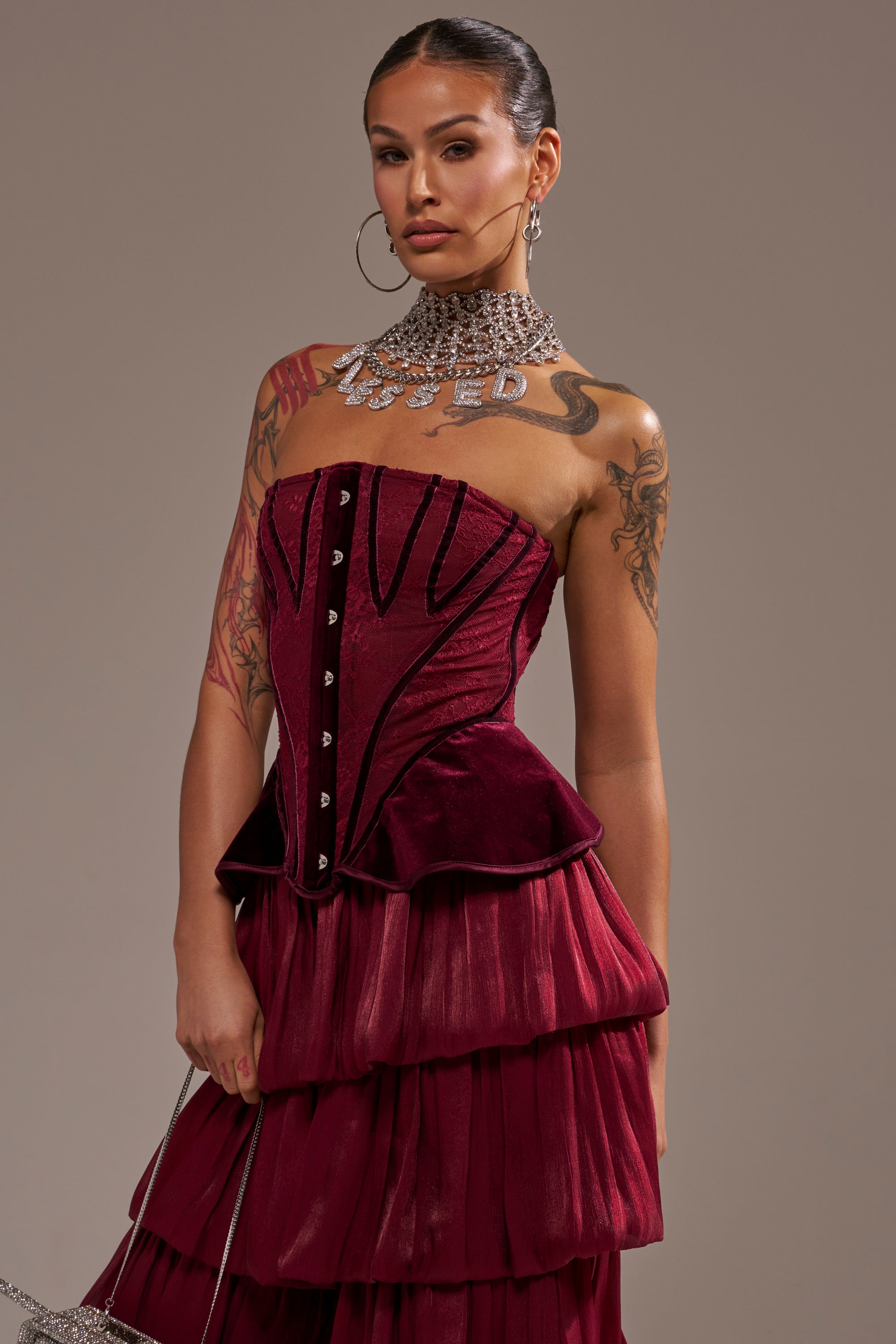 A tattooed woman wears the RUBY ROSE CORSET in strapless burgundy velvet with sheer ruffles, styled with a bold silver necklace and hoop earrings. She poses confidently against a plain background, looking at the camera. 0