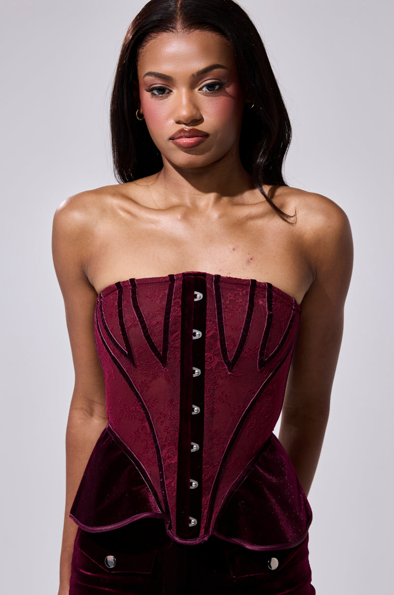 A woman with long dark hair models the strapless RUBY ROSE CORSET, featuring velvet panels, decorative buttons, and black trim, while standing against a plain light gray background. 2