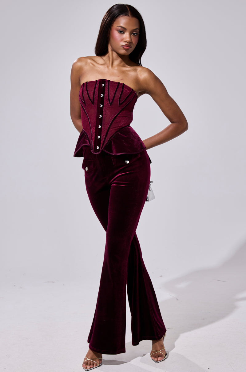 A woman poses in the AKIRA Label RUBY ROSE CORSET, a strapless velvet-paneled top with decorative boning, paired with matching flared velvet pants and strappy heels against a plain light background. 6