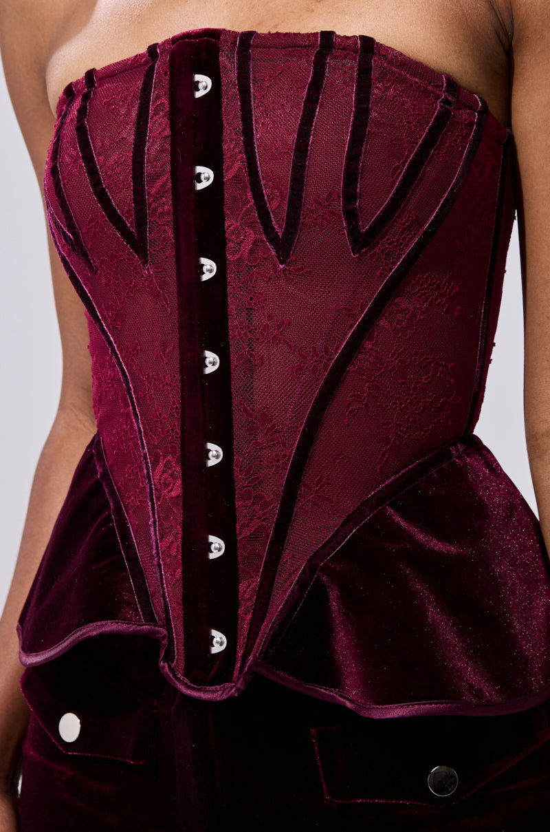 A close-up of a person wearing the RUBY ROSE CORSET, a strapless deep burgundy velvet-paneled piece with sheer lace details, vertical boning, front decorative buttons, and a peplum-style waist flare. 7