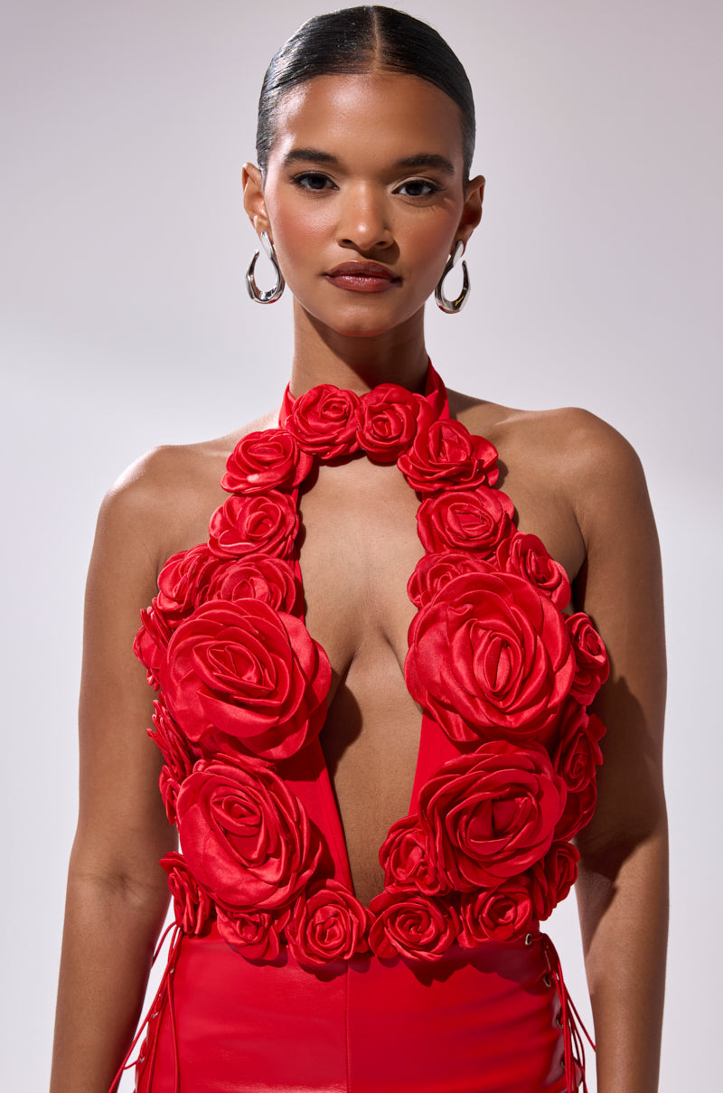 A woman with slicked-back hair and hoop earrings models the GIVE ME A ROSE BODYSUIT, a bold red one-piece with 3D satin rose appliques and a deep plunging neckline, posing confidently against a plain light background. 0
