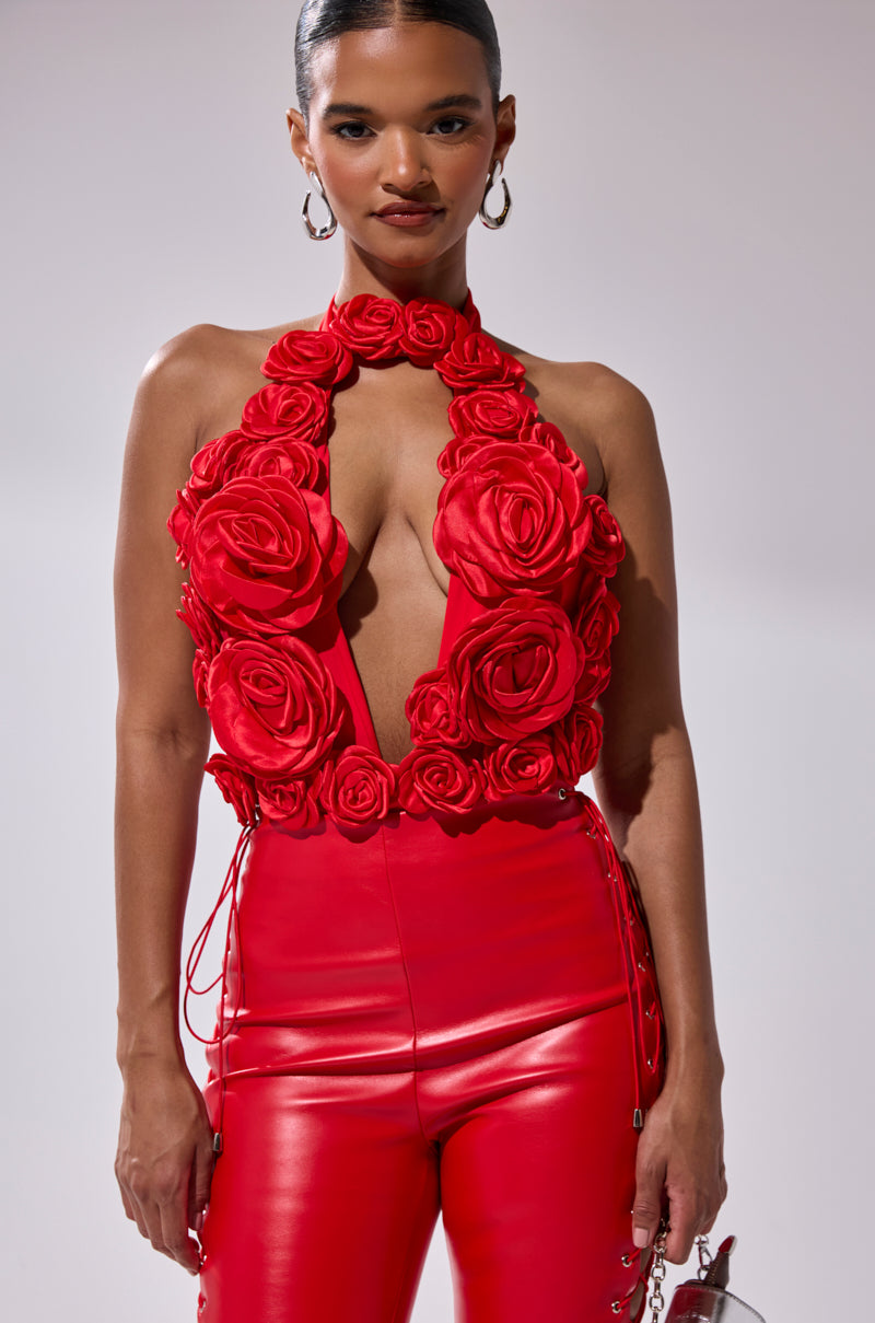 A woman wears the GIVE ME A ROSE BODYSUIT featuring bold red with a plunge halter neckline and large 3D satin rose appliques, styled with slicked-back hair, hoop earrings, and a small metallic bag. 1