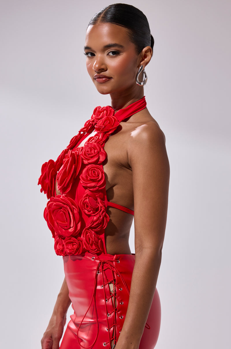 A woman wears the GIVE ME A ROSE BODYSUIT, a bold red piece with a plunge halter neckline, large fabric flowers, and a fitted skirt. She pairs it with sleek hair and silver hoop earrings against a light background. 2