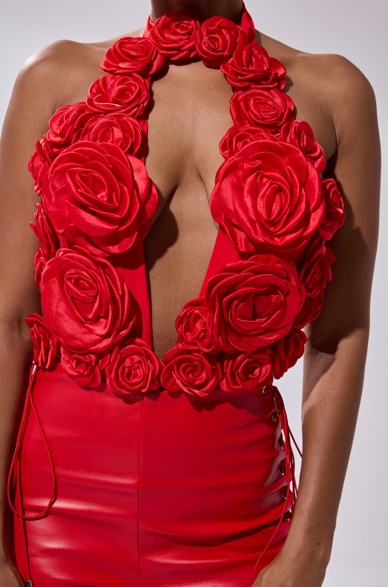 A person wears the GIVE ME A ROSE BODYSUIT, a bold, sleeveless red piece featuring a plunge halter neckline and large 3D satin rose appliques on the bodice. The shiny skirt has lace-up sides. The backdrop is plain light gray. 5