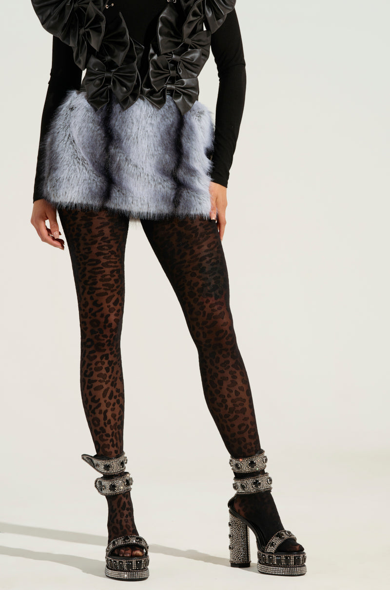 A person dressed in an eye-catching fashion ensemble that includes a black top with dramatic ruffles, the standout ICE QUEEN FUR MINI SKIRT, leopard print tights, and high-heeled sandals adorned with sparkling details poses against a simple background.