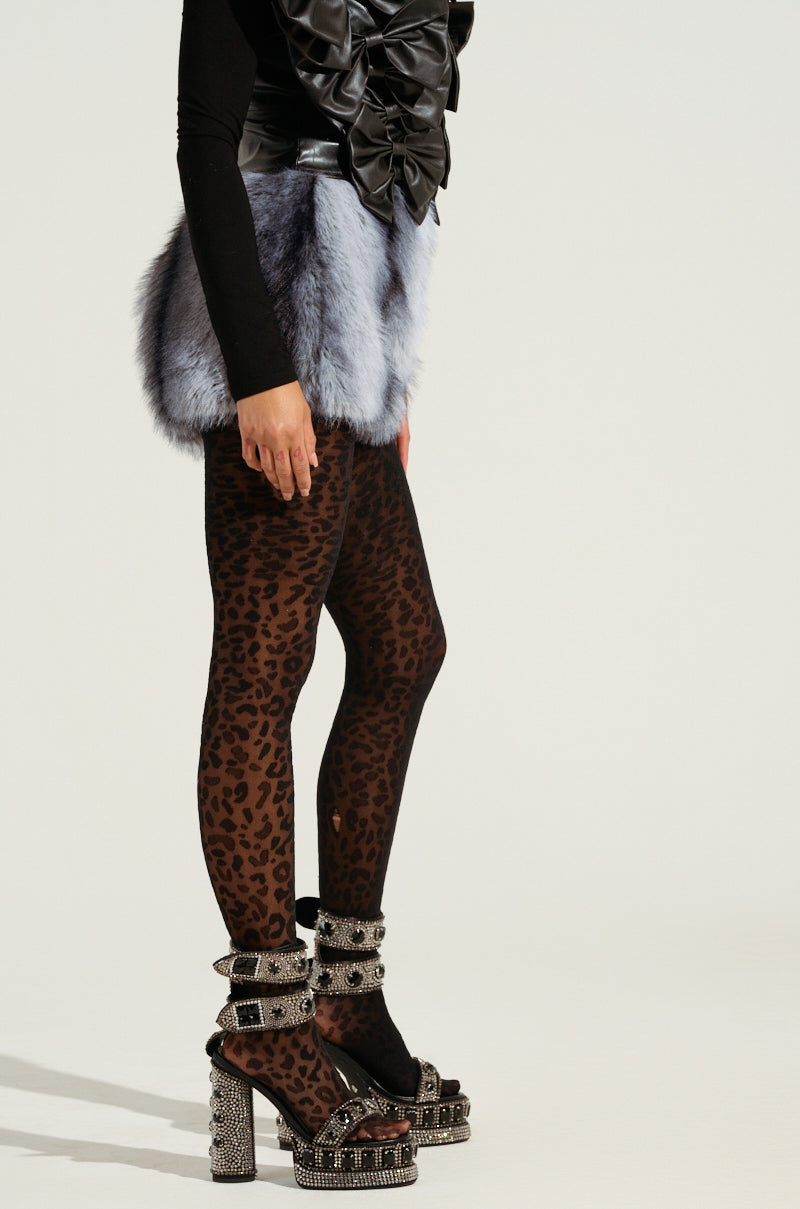 The individual is dressed in a black top featuring prominent ruffles and the stunning ICE QUEEN FUR MINI SKIRT, complemented by leopard print leggings. They complete the ensemble with high-heeled platform sandals embellished with studs and rhinestones, all displayed against a simple off-white backdrop.