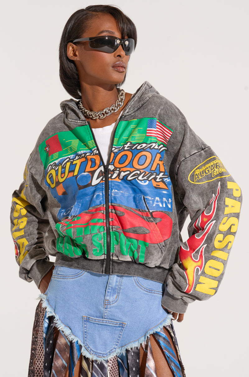 A stylish person wearing sunglasses stands confidently in the OFF TO THE RACES GRAPHIC ZIP UP HOODIE, adorned with vibrant racing-inspired graphics and patches like "Outdoor," "Sports," and "Passion." They also wear a blue denim skirt with a patchwork design.