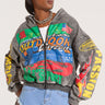 A stylish person wearing sunglasses stands confidently in the OFF TO THE RACES GRAPHIC ZIP UP HOODIE, adorned with vibrant racing-inspired graphics and patches like "Outdoor," "Sports," and "Passion." They also wear a blue denim skirt with a patchwork design.