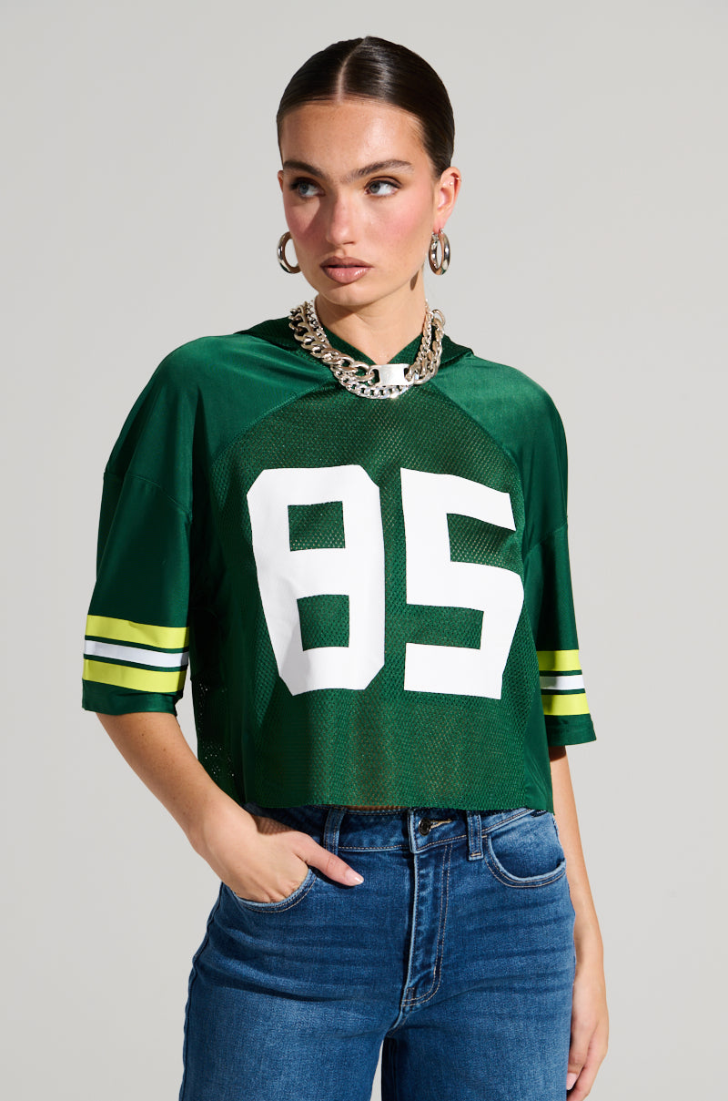A person with dark hair tied back is wearing the LUCKY JERSEY, a green semi-stretchy jersey material top featuring the number 85 and striped sleeves, paired with a chunky chain necklace. They have one hand in the pocket of their blue jeans and are glancing to the side against a plain background.