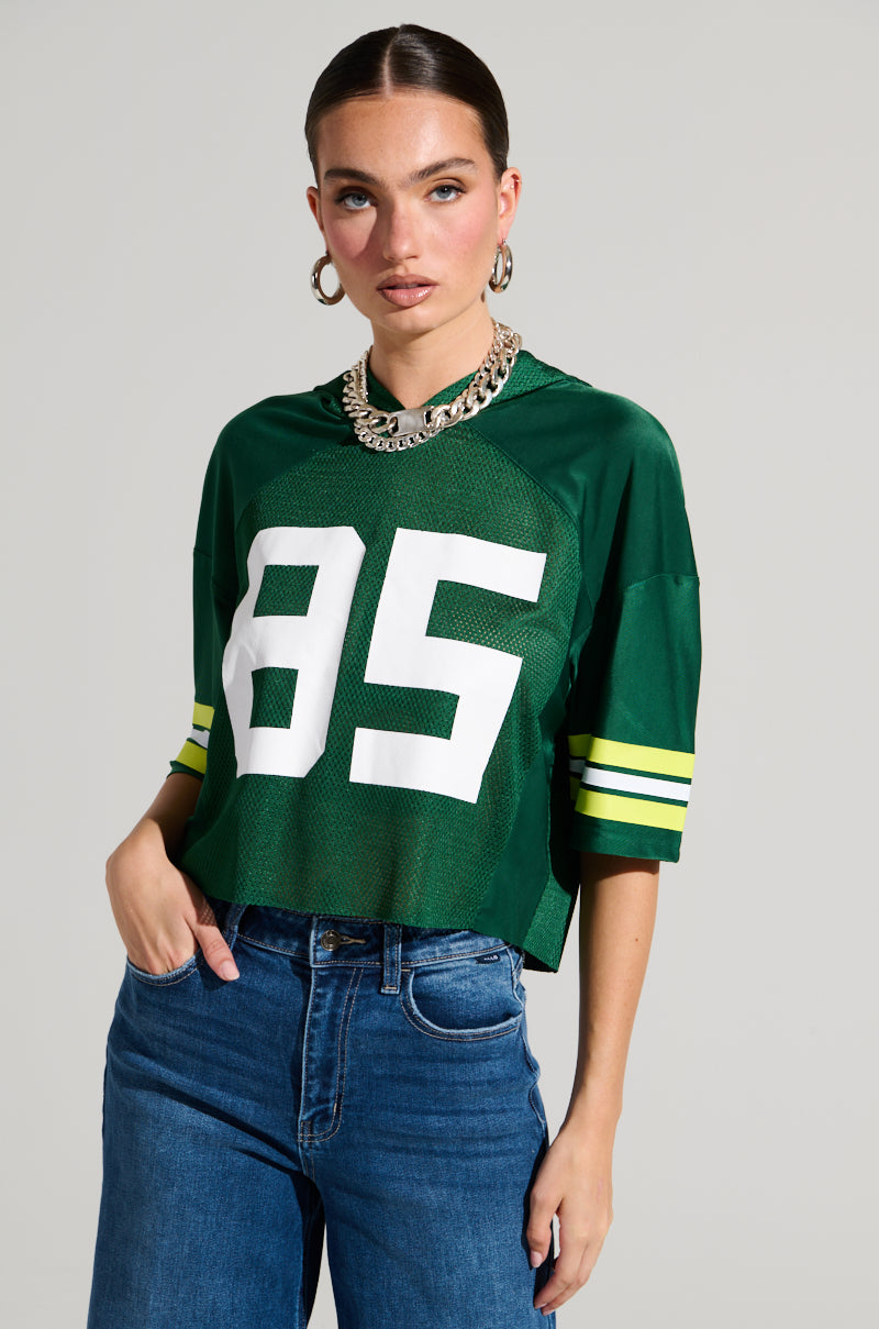 A person with slick, pulled-back hair is wearing a green LUCKY JERSEY made of semi-stretchy material, featuring the number 85 and yellow striped sleeves. They pair it with blue jeans, large hoop earrings, and a chunky silver necklace against a plain, light-colored wall — their sporty chic top completes the look.